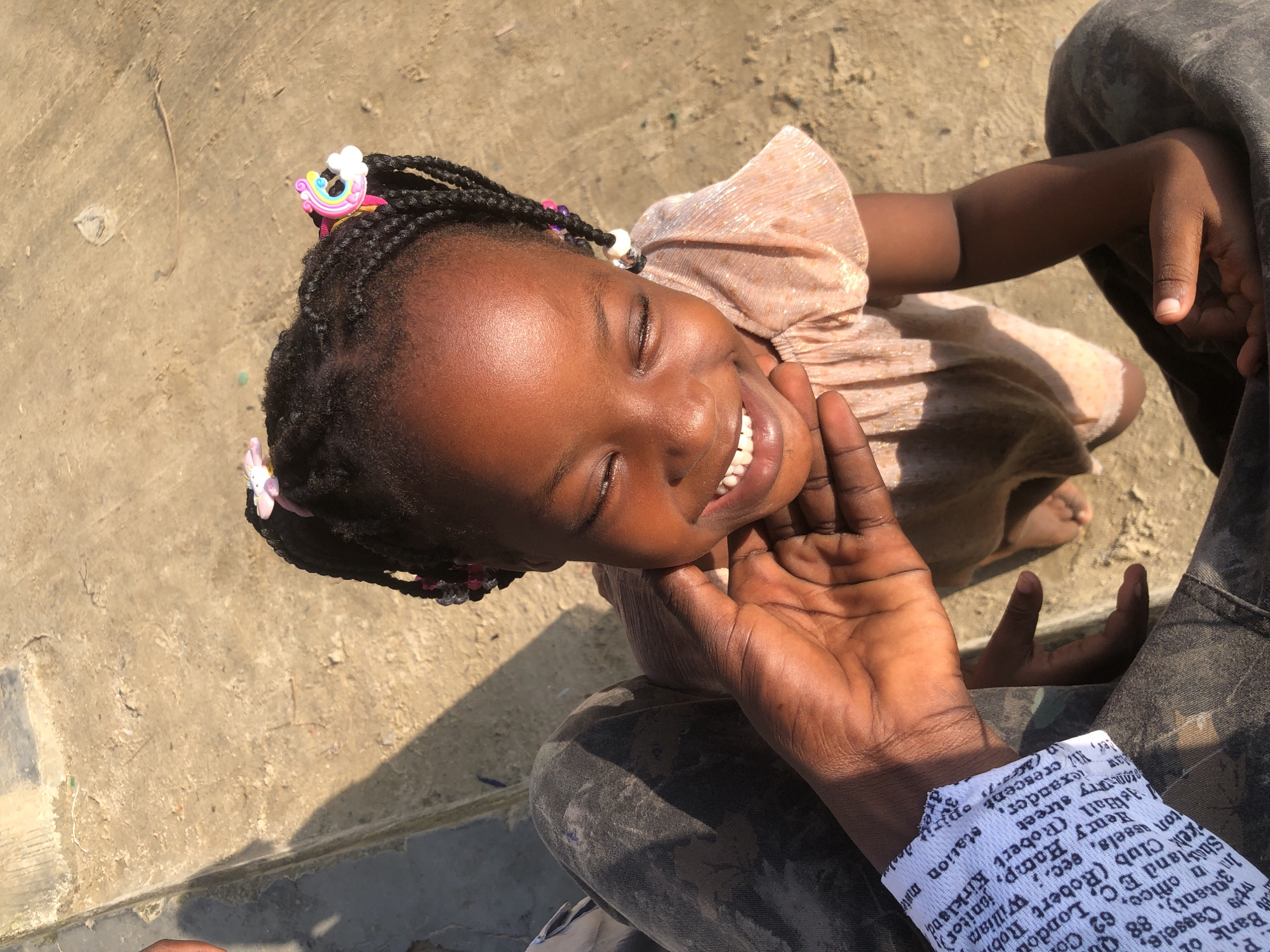 A high-angle, close-up shot shows a young Black girl in Ado, Nigeria, beaming with joy as someone's hand gently supports her chin. The girl's eyes are closed in a smile, and her lips are parted, revealing white teeth. Her dark hair is styled in braids, adorned with colorful hair clips shaped like rainbows and flowers, and small beads. She wears a shimmery, light-pink dress with ruffled sleeves. The hand supporting her chin is darker skinned, and the person's forearm, visible on the left, is covered in camouflage-patterned pants and a white sleeve with black text. The background is a rough, concrete surface bathed in sunlight, casting a long shadow. The scene suggests a warm, outdoor setting in a city.
