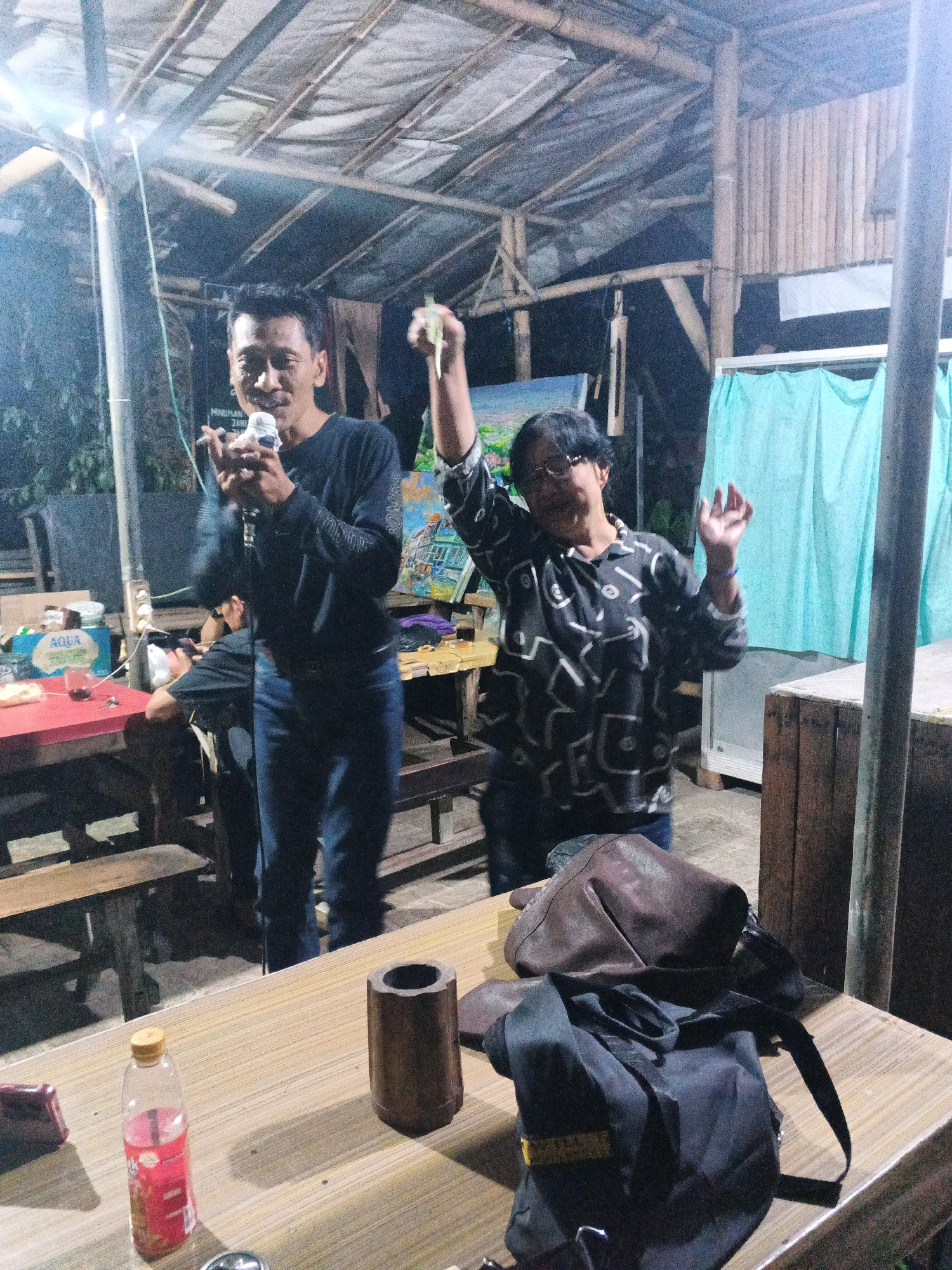 The image depicts an outdoor gathering at night, likely in Ungaran, Indonesia, under a makeshift roof supported by bamboo and wood. Two individuals are prominent in the foreground. A man on the left, wearing a black long-sleeved shirt and blue jeans, is holding a microphone and appears to be singing or speaking. To his right, a woman in a dark patterned shirt and dark pants is enthusiastically gesturing with her hands in the air, seemingly enjoying the event.
In the background, tables with chairs are visible, along with some bottles and a glass on a red table on the left, suggesting a casual dining or drinking area. A blue curtain hangs on the right side of the scene. The overall atmosphere appears to be lively and informal. In the immediate foreground, a wooden table displays a plastic bottle of red liquid, a cylindrical wooden object, and two bags – one brown leather and one black fabric with a strap. The lighting is dim, with artificial lights illuminating the scene.