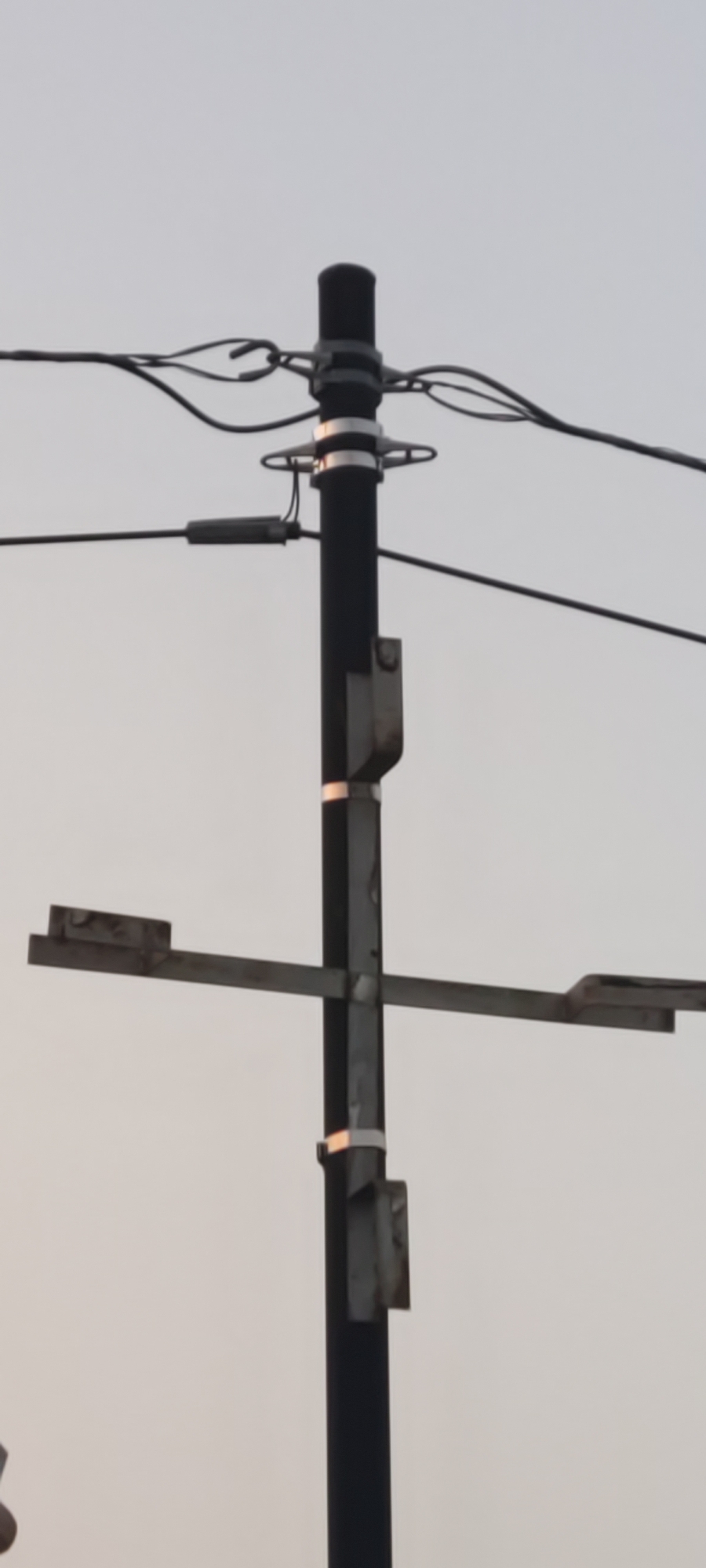 The image shows a utility pole with electrical wires against a pale sky. Attached to the pole are horizontal metal brackets, suggesting the presence of streetlights or other attachments. The pole itself is dark and cylindrical, with metal bands wrapped around it at intervals. One of the wires appears to have a gray cylindrical device attached to it. The scene is captured from a low angle, looking upwards at the pole and wires. This is a typical view of infrastructure in Pakusamben, Indonesia, where such poles are common for power distribution and street lighting.