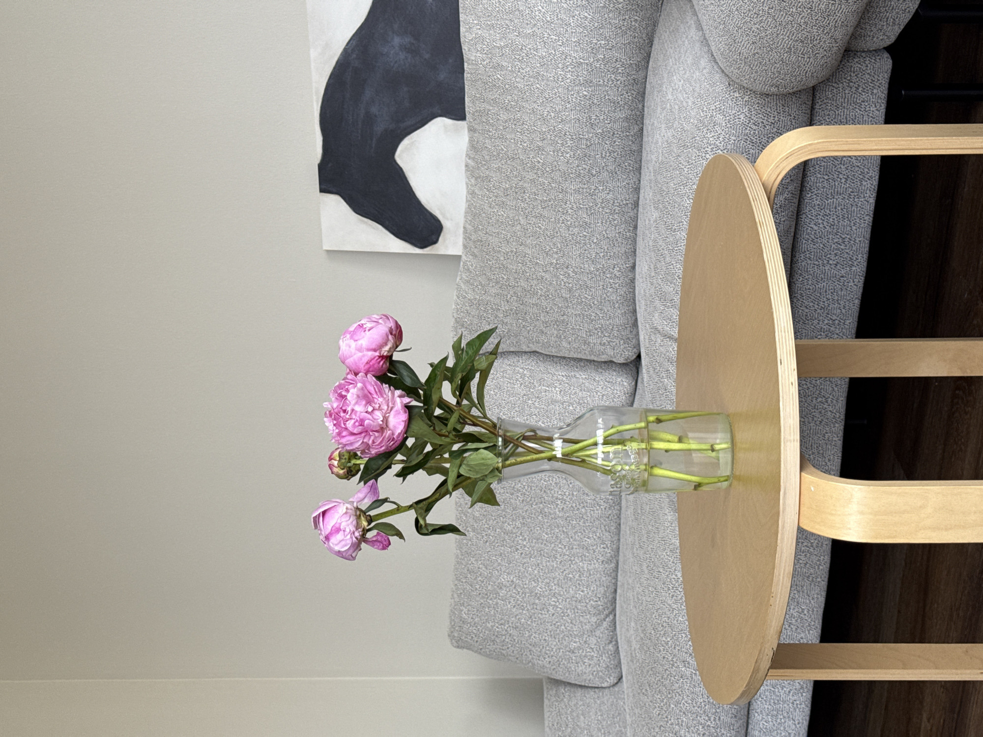 The image displays a still life arrangement in a domestic setting. The primary subjects are a vase of pink peonies placed on a wooden side table, positioned next to a grey sofa. Above the sofa, a portion of a large black and white abstract painting is visible on a light grey wall. The sofa appears to be made of a textured fabric, with visible cushions. The wooden side table has a rounded top and a minimalist design with curved wooden supports. The vase holding the flowers is made of clear glass and contains water with visible stems. The peonies themselves are a vibrant pink, with some buds still closed. The lighting suggests it is daytime, likely indoors, with a soft, diffused light. The overall mood is calm and serene, suggesting a well-decorated living space. There is no visible text in the image. The context provided places this scene in Chicago, United States of America.