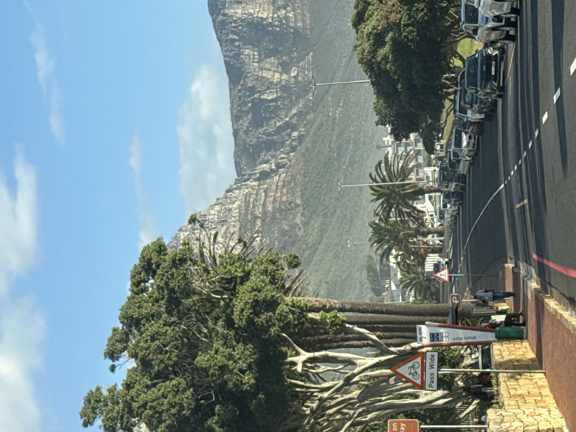 The image shows a street in Cape Town, South Africa, with a view of Lion's Head Mountain. The street is lined with parked cars and trees. On the right side, a sidewalk with a brick wall is visible. There are street signs, including a "Pass Wide" sign for cyclists, as well as a street advertisement sign. A couple of people are walking along the sidewalk. The sky is blue with some white clouds. The mountain occupies a significant portion of the background.