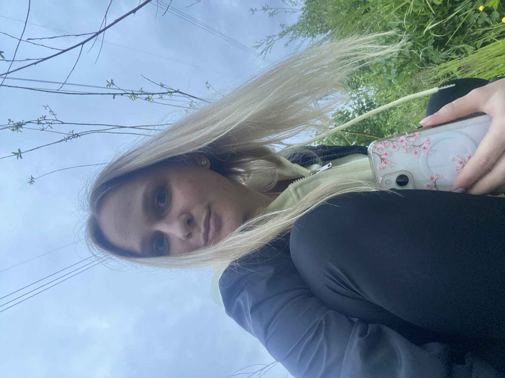 The image displays a young adult female captured from a low-angle perspective in an outdoor setting under an overcast sky. She has long, straight blonde hair, a fair complexion, and is looking directly at the camera with a neutral expression. She wears a light green-yellow hooded sweatshirt with a zipper, layered under a dark blue or black jacket. Dark leggings or pants are visible on her lower body. A small, clear stud earring is present in her visible right ear. Her right hand, positioned in the lower right corner, holds a smartphone with a clear case featuring pink cherry blossom designs and a visible white MagSafe ring. The background consists of a pale blue-grey sky with faint clouds, crossed diagonally by multiple thin power lines or wires. Various green vegetation, including tall grass and thin tree branches with emerging leaves, is present in the mid-ground and background. The environment appears to be an unspecific natural or semi-urban outdoor area, potentially a park or undeveloped land in the city of Unknown, Unknown.