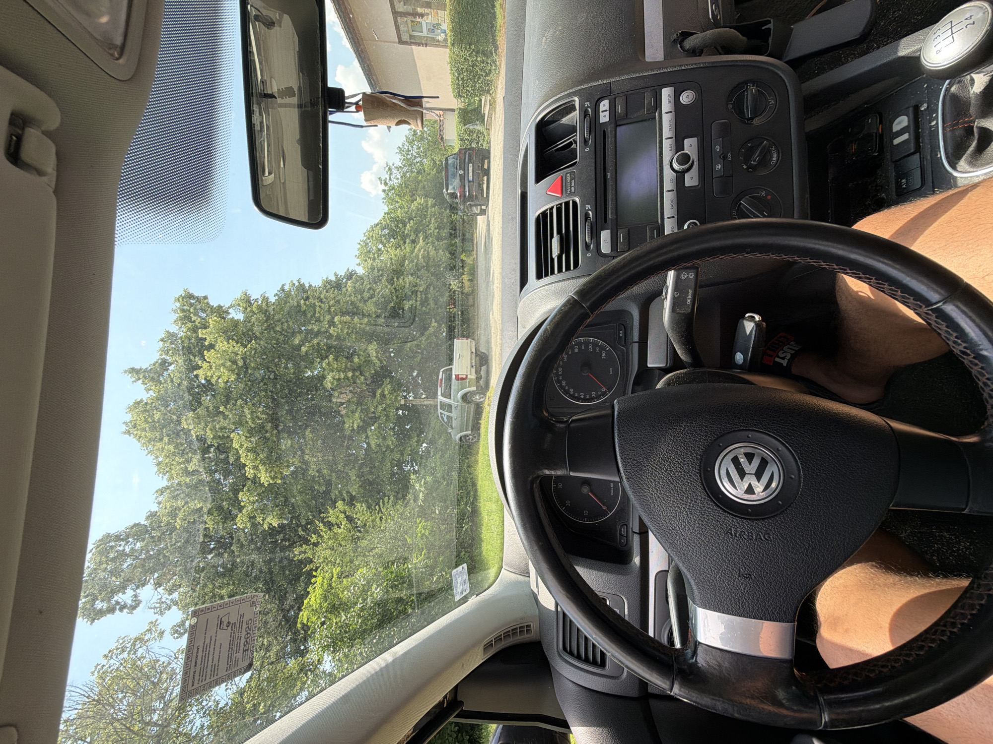 The image presents a first-person view from the driver's seat of a parked Volkswagen Golf Mk5, characterized by its steering wheel with the VW logo, a dashboard displaying a speedometer (up to 260 km/h) and tachometer, and a center console with infotainment system controls and manual climate dials. The vehicle has a manual 6-speed transmission with a black leather shift boot featuring red stitching. Keys, one with a "JUST" keychain, are in the ignition. The driver's bare thighs, clad in shorts, are visible in the lower foreground. A small, wooden, bird-shaped charm hangs from the rearview mirror. A vignette or inspection sticker on the upper left of the windshield contains text in German and English, notably "WICHTIG / IMPORTANT!" and the year "2025."
The vehicle is stationary on an unpaved, dirt and gravel parking area. Directly ahead, a silver/light green pickup truck, likely a Great Wall Wingle, is parked facing away from the camera. Further down the path, a dark-colored SUV, identified as a Volkswagen Touareg, is also parked facing away, displaying a Bulgarian license plate "P H 7523 H P." The surrounding landscape is dominated by numerous large, leafy green trees, suggesting a lush, natural environment. On the far right, a light-colored building with a tiled roof is partially visible. The sky is clear blue with some scattered white clouds, indicating bright, sunny weather. The scene is located in Dabene, Bulgaria, consistent with the Plovdiv Province license plate.