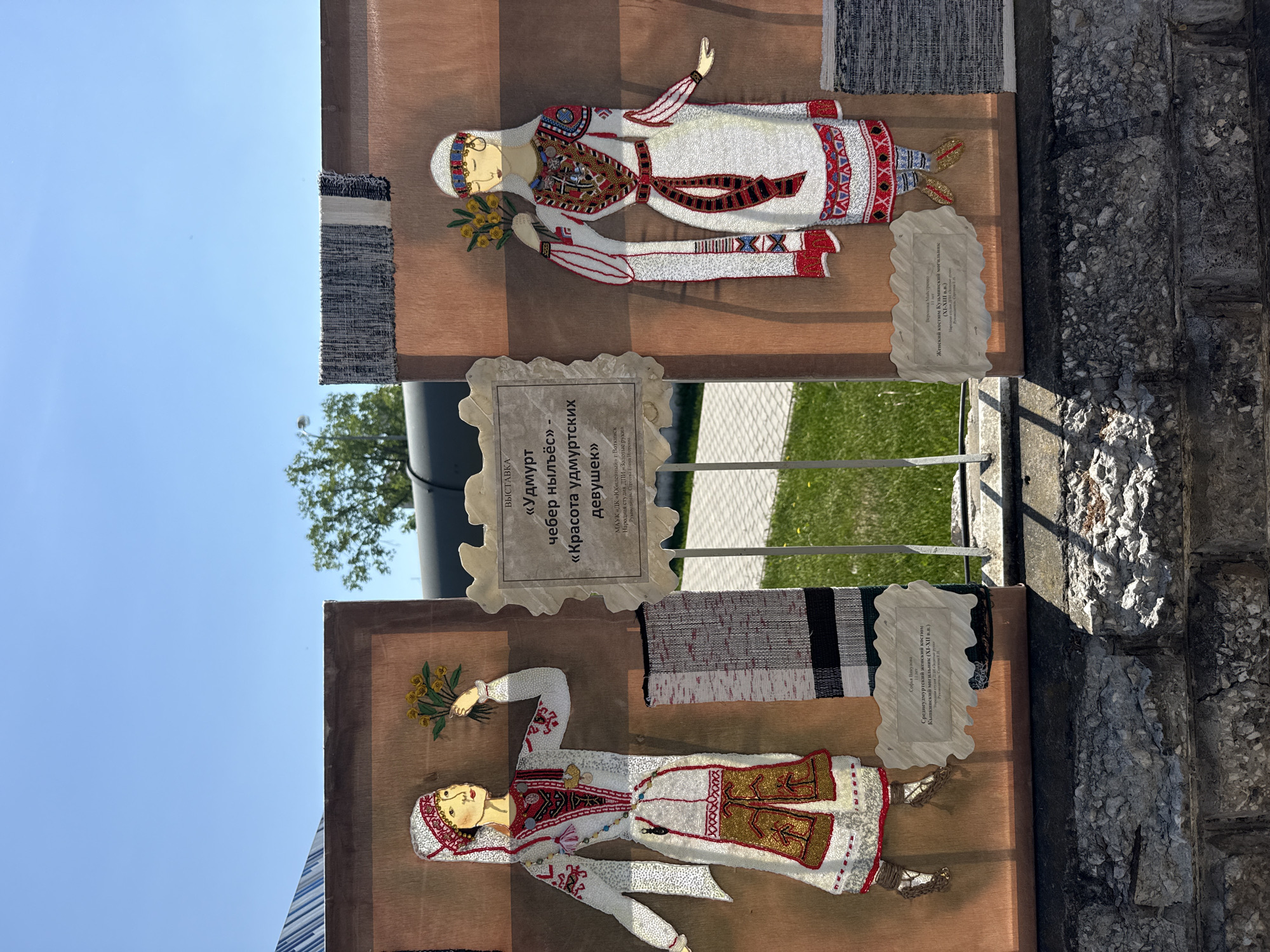 The image shows an outdoor display in Votkinsk, Russia, featuring folk art panels depicting women in traditional Udmurt costumes. Two panels are visible, mounted vertically, each with a beaded representation of a woman in white clothing with red and gold accents. Each panel also has a woven textile strip attached at the top. Between the panels is a sign that says "Udmurt cheber nyljos - Krasota Udmurtskikh devushek" which translates to "Udmurt Beauty Girls". One panel cites the costume as a "Sredneudmurtskiy zhenskiy kostyum", or "Central Udmurt Women's Costume". Another indicates a "Zhenskiy kostyum Kuzminskiy mogilnik", or "Women's Costume Kuzminskiy Burial Ground". The panels are displayed outside on a bright, sunny day near a stone wall or structure, with grass visible in the background. The sign indicates the display is part of an exhibit by the "DK Yubileyny" (House of Culture Yubileyny) of Votkinsk, specifically the "Zolotye Ruki" (Golden Hands) folk art studio led by Elena Igorevna Sergeeva.
