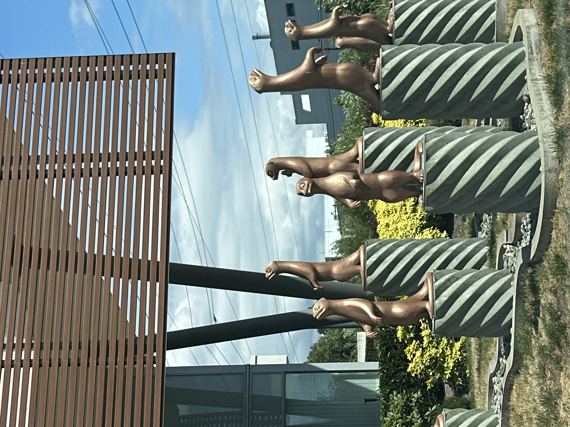 In Burnaby, Canada, a bronze sculpture of otters stands on twisted pedestals outside a modern building. The otters, depicted in various playful poses, are arranged in a line. The building features a glass facade and a slatted wooden overhang. The surrounding environment includes green shrubbery, yellow flowering plants, and a cloudy sky with power lines visible overhead. The scene appears to be a public art installation or part of an architectural design.