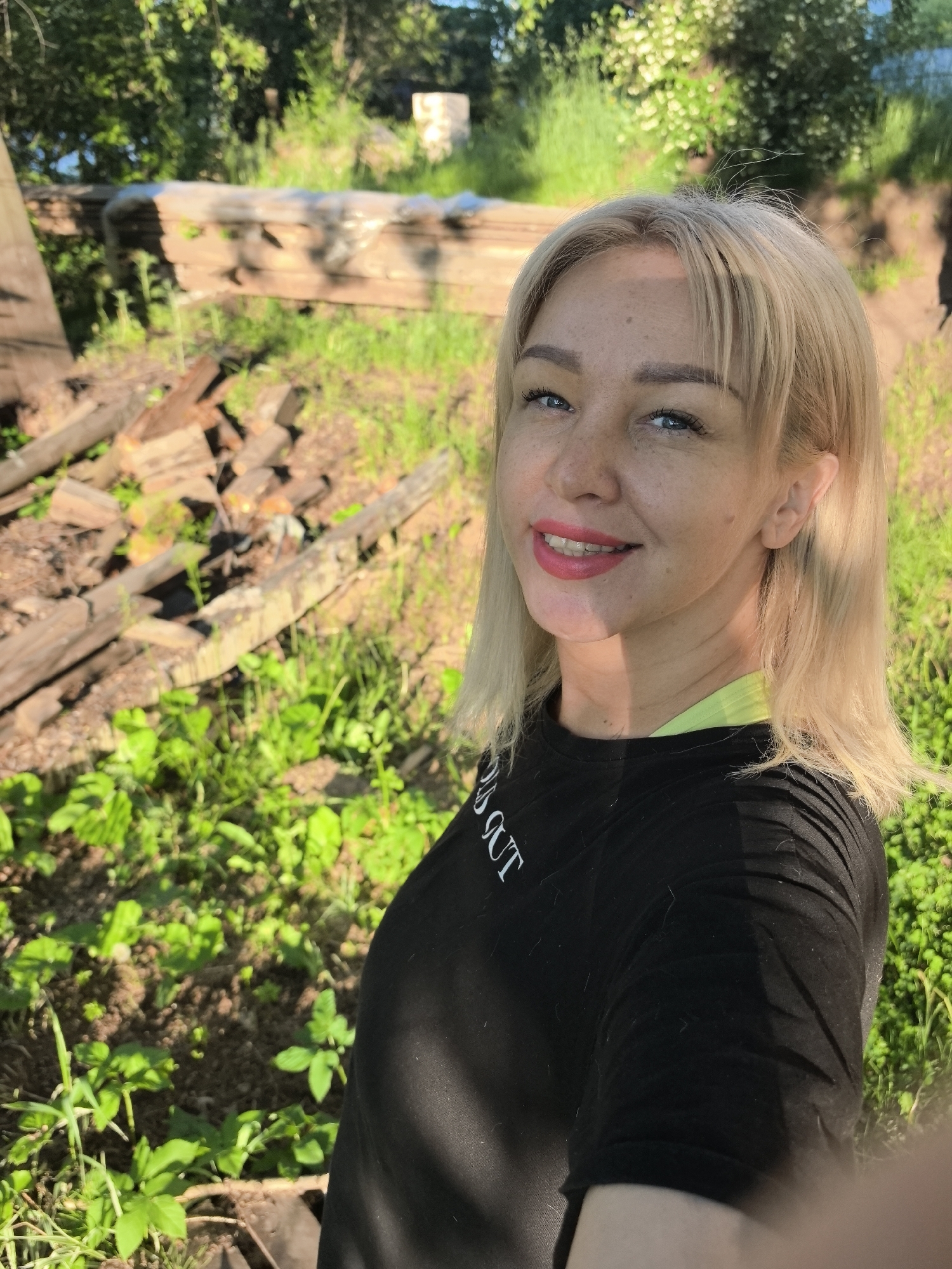An outdoor, eye-level selfie shows a blonde female in the city of Noviy, Russia. She has blue eyes, a freckled complexion, and is smiling, revealing upper teeth with bright pink lipstick. Her blonde hair is shoulder-length with bangs. She is wearing a black short-sleeved t-shirt with partially visible white text on the left shoulder and a neon green garment underneath, visible at the collar. Strong sunlight illuminates her face and the scene, casting shadows on her neck and the left side of her body. The background consists of green foliage, including grass and broad-leafed plants, on uneven soil. To her left and behind, a pile of cut wooden logs and scattered timber is visible. Further back, horizontally stacked wooden planks, wrapped in a translucent material, extend across the mid-ground. In the far background, partially obscured by trees and bushes, a light-colored, possibly concrete, cylindrical or rectangular structure stands on the right.