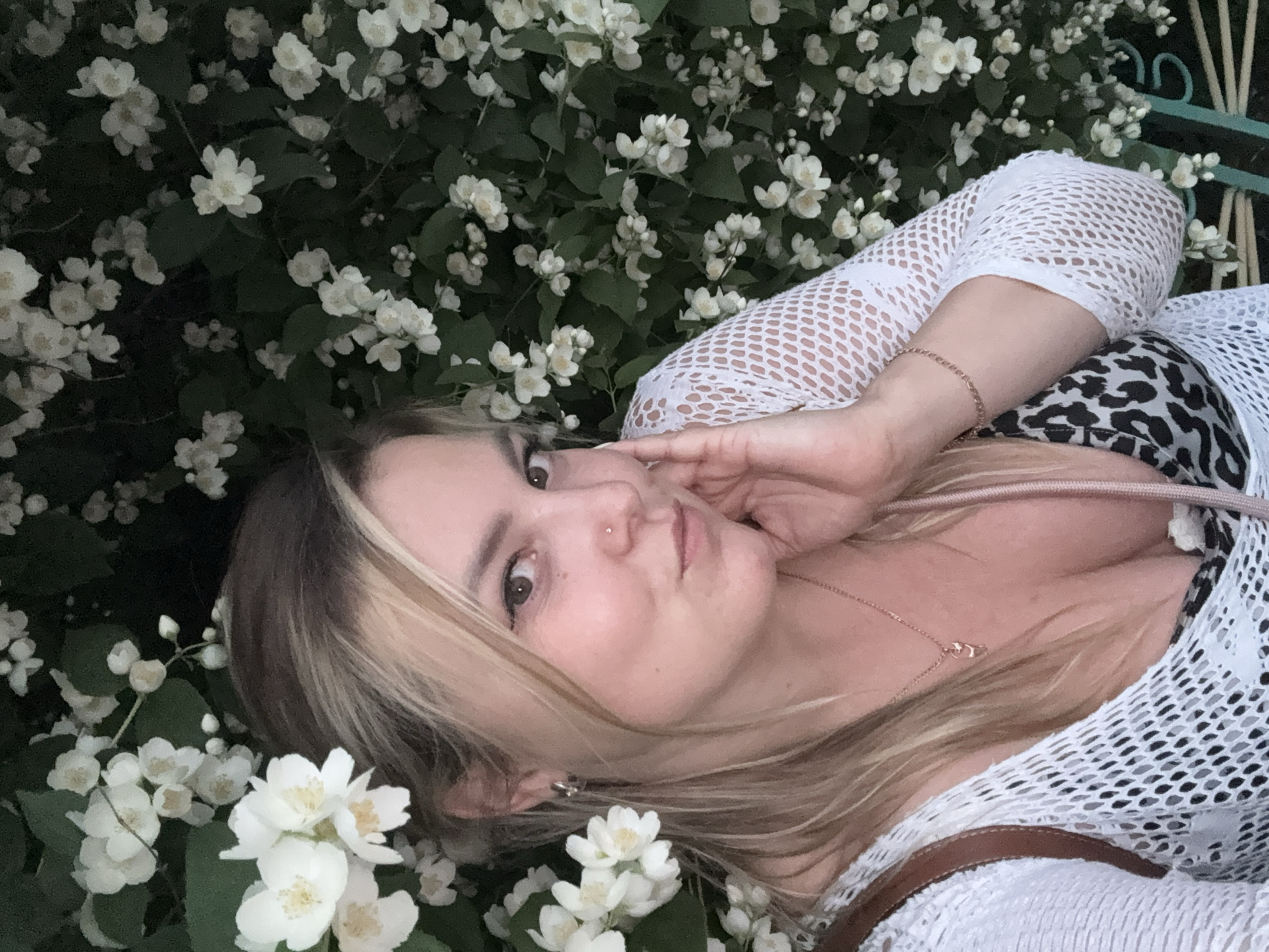 A woman is posing for a selfie in front of a dense bush of white jasmine flowers. She has blonde hair, is wearing a white mesh top over a leopard print garment, and a rose gold necklace. Her left hand is gently touching her face. The image is a medium shot and focuses on the woman and the flowers. The background is dark, with the jasmine flowers providing the only significant light. The scene appears to be outdoors, possibly in a garden or park setting within the city of Chaikovsky, Russia.