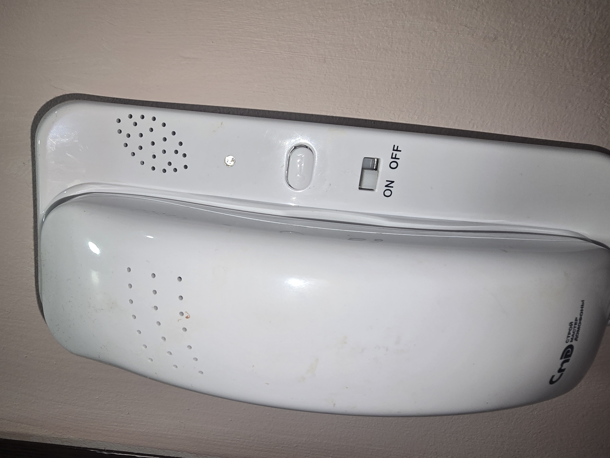 The image displays a white intercom handset mounted on a light pink wall. The handset is oriented vertically. It features several perforated speaker/microphone areas, a push-button, and a toggle switch labeled "ON OFF." Below the switch, the Russian text "СТРОЙ МАСТЕР ДОМОФОНЫ" is visible, indicating the manufacturer or brand, likely translating to "Stroy Master Intercoms." A small LED light is also present above the push-button. The wall appears to be textured and a darker vertical line is visible on the left edge of the frame, suggesting a corner or doorway. No people or specific actions are depicted. The overall scene is static and domestic or commercial, consistent with an apartment building hallway or entrance area. The location being Ulan-Ude, Russia, is stated as context but not visually discernible from the image itself.