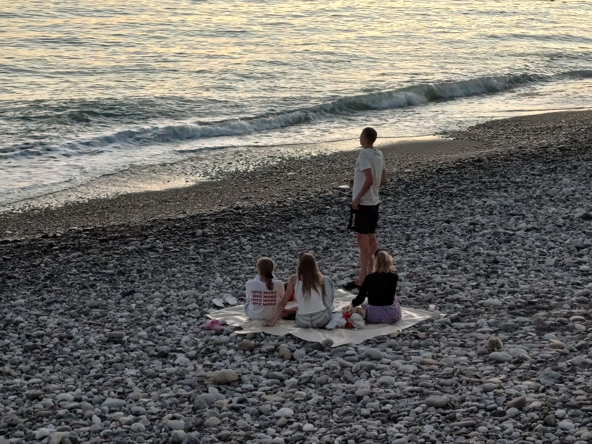 In Sochi, Russia, on a pebbled beach, a group of four individuals are gathered. Three young females are seated on a light-colored blanket, facing the sea. One of them wears a white sweatshirt with red lettering that reads "LOVE MORE LOVE MORE LOVE MORE LOVE MORE". A fourth individual, a male, stands further back on the beach, facing the sea. The sea is calm with small waves lapping at the shore. The lighting suggests it is either early morning or late evening.