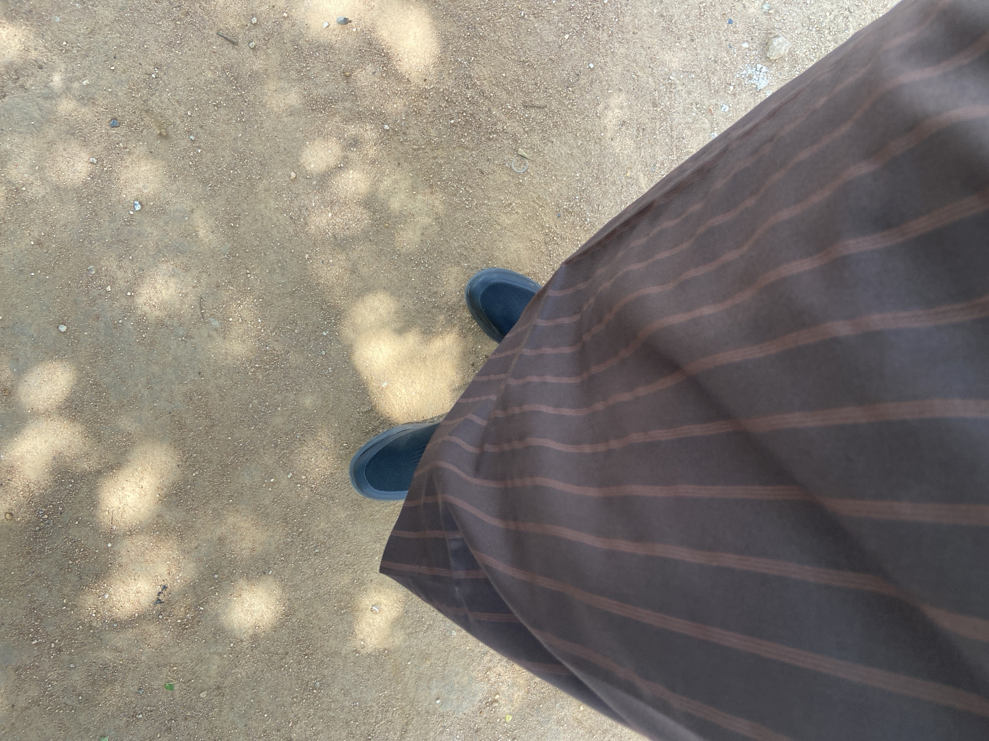 An overhead shot shows a person's legs and feet, wearing dark blue slip-on shoes and a brown striped skirt, standing on a dirt path dappled with sunlight. The skirt is draped over the person's left leg, with vertical brown stripes on a darker brown fabric. The ground is composed of fine, light brown dirt with small pebbles and scattered leaves, illuminated by patches of sunlight and shadow.