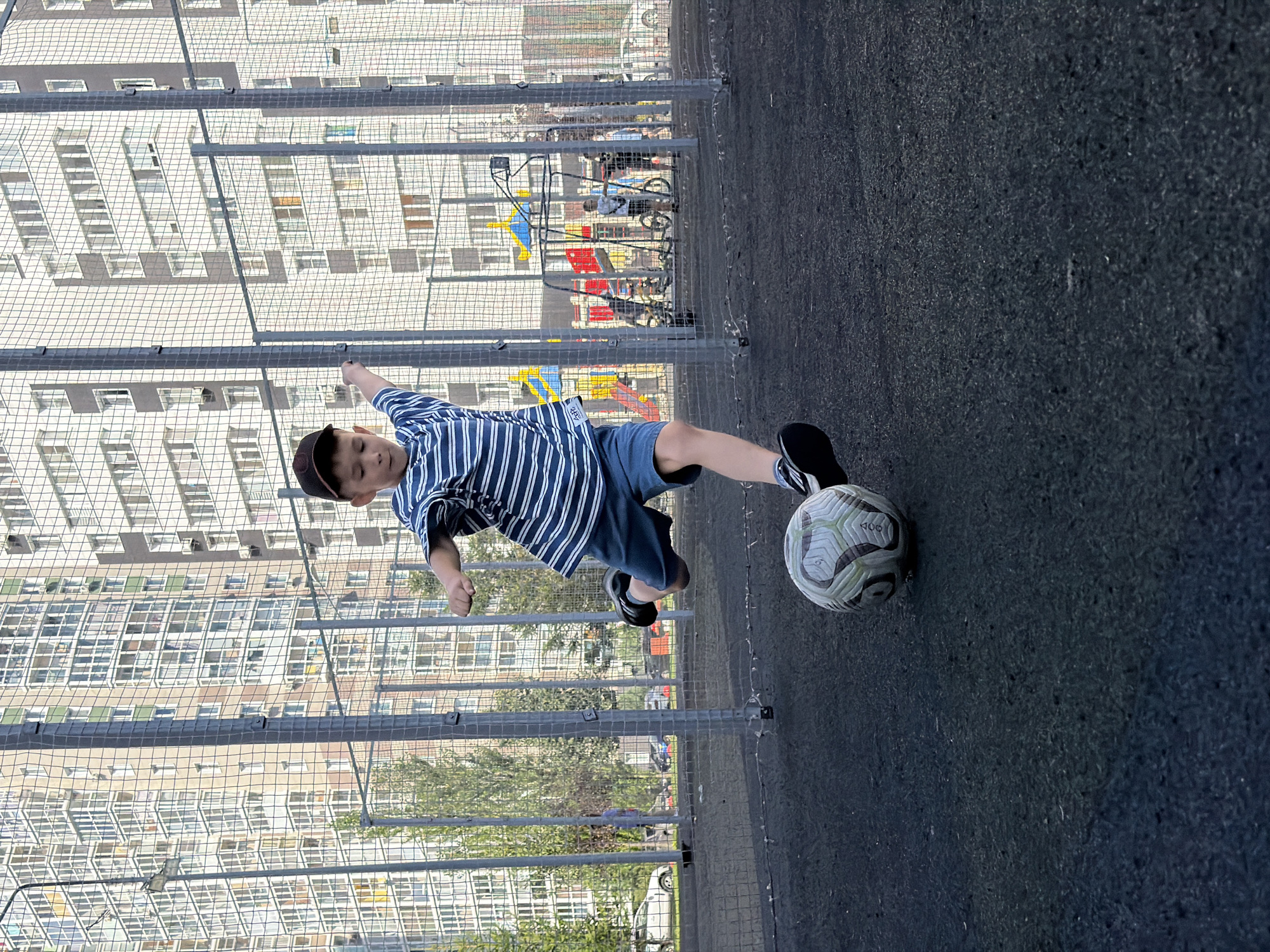 A young boy wearing a striped t-shirt, blue shorts, and a baseball cap is kicking a soccer ball on an outdoor sports court. The court has a dark, textured surface and is surrounded by a tall chain-link fence. In the background, there are modern apartment buildings with many windows, trees, and a playground with colorful equipment. Several other people, including children and adults, are visible in the background, some on bicycles. The scene appears to be taking place in a residential area of Kazan, Russia, likely a public recreational space.