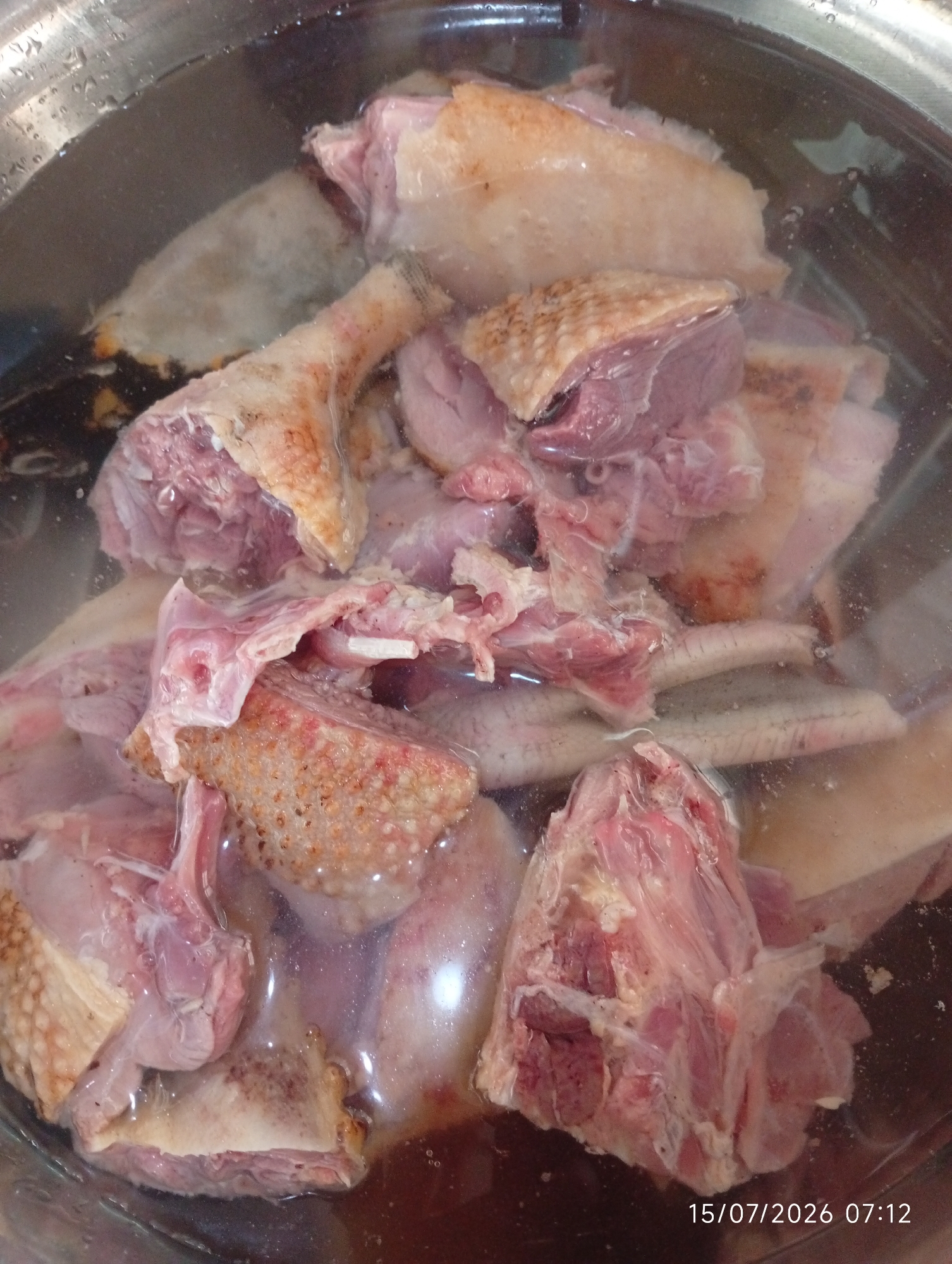 In a metal pot filled with water, several pieces of raw duck meat are submerged. The duck pieces appear to be jointed, with visible skin and bone structures. The water is slightly cloudy, with some darker areas suggesting impurities or cooking liquids. Bubbles are present on the surface of the water and on the sides of the pot, indicating recent activity or the start of a cooking process. The lighting in the image is direct, casting reflections on the water and highlighting the textures of the meat and the pot. The timestamp "15/07/2026 07:12" is visible in the lower right corner, suggesting the image was captured on that date and time. The scene is consistent with food preparation, specifically the initial stages of cooking duck. The location is Bana Stream, Nigeria, which implies a domestic or possibly a small-scale commercial food preparation setting within that area. No people are visible in the image.