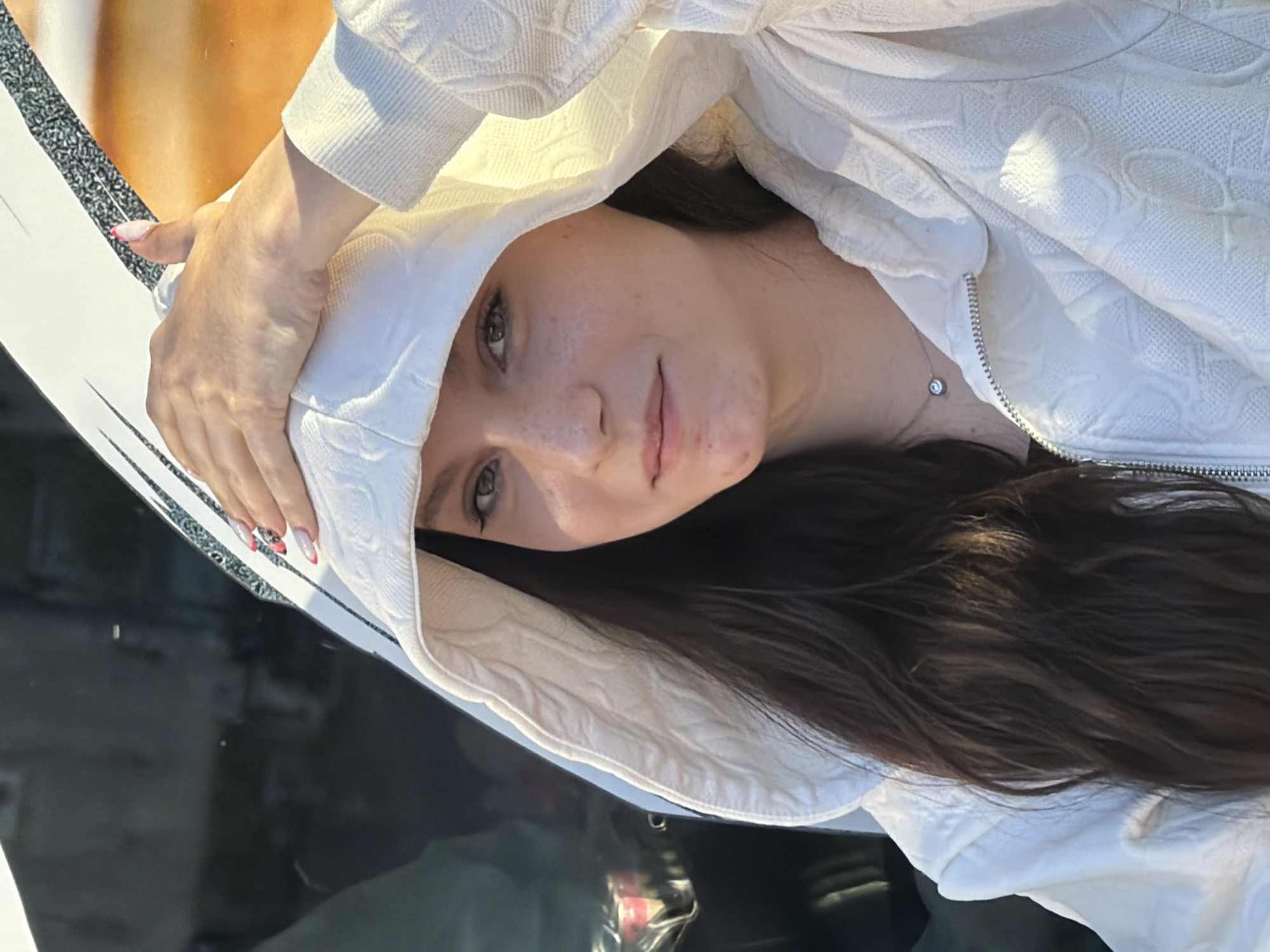 The image is a portrait of a young woman, rotated ninety degrees counter-clockwise. When viewed correctly, she is looking directly at the camera with a subtle, serene expression. She has fair skin, dark eyes, and long, dark brown hair that cascades over her right shoulder. She is wearing a white hooded jacket or hoodie, which features a distinctive quilted or embossed pattern of repeating circular or 'B' shapes. A silver zipper is visible on the jacket. Around her neck, a delicate silver necklace with a small, clear gemstone pendant can be seen.
Her left hand is raised, gently holding the edge of her hood near her temple, with the sleeve of the jacket visible. Her fingernails are adorned with a light pink base and intricate red and black designs on the tips. The bright lighting suggests it is daytime, likely sunny, as indicated by the highlights on her clothing and skin.
The background directly behind her head and upper body shows an out-of-focus blend of warm orange and brown tones, possibly suggesting a building facade or natural landscape. Below her, there is a dark, highly reflective surface, likely the hood of a car or a similar glossy object, reflecting faint hints of the sky and surroundings. The scene depicts a casual outdoor setting in Shumen, Bulgaria. No visible text is present in the image.