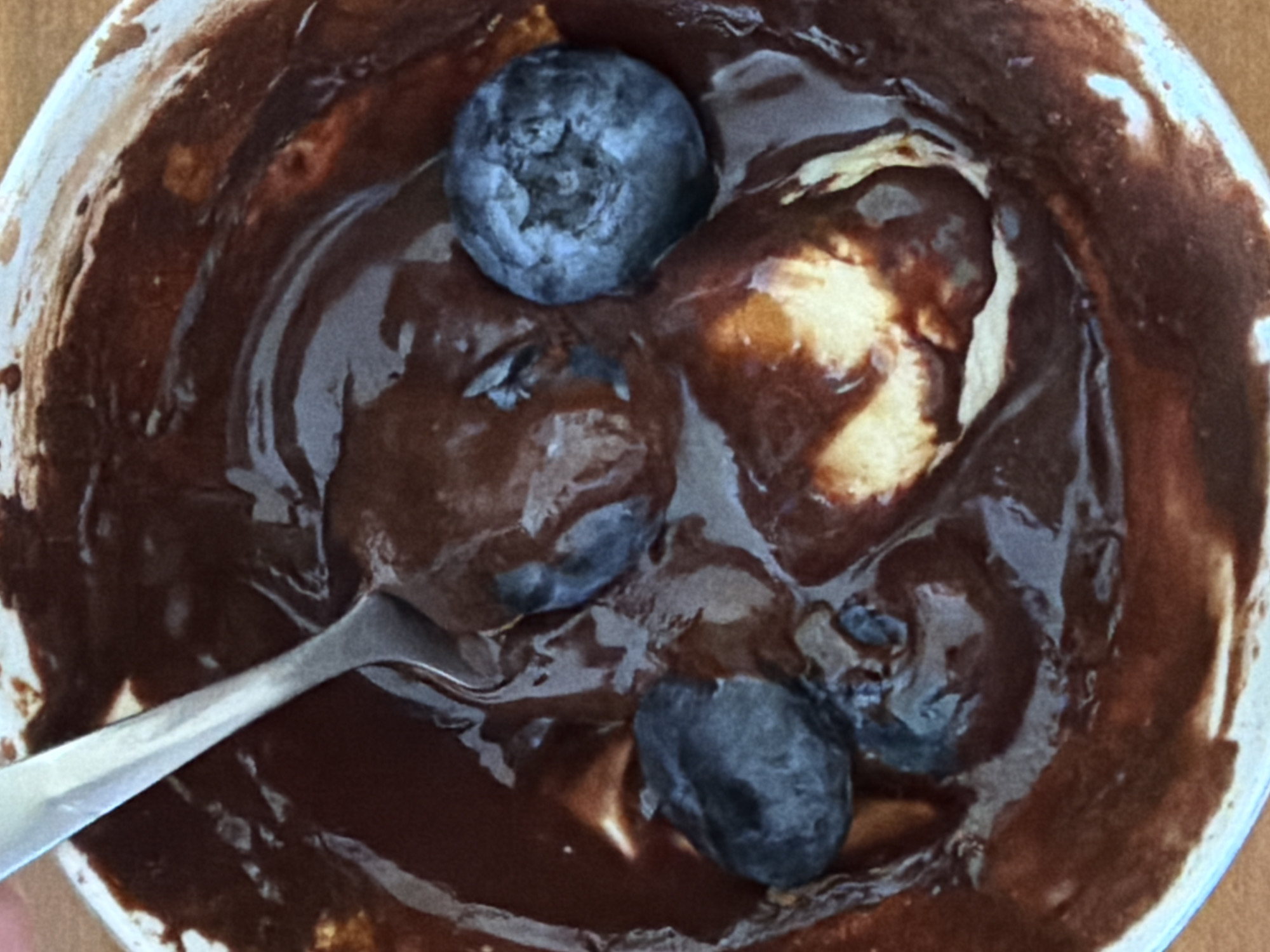 This image depicts a close-up, top-down view of a dessert contained within a white bowl, located in the city of Khimki, Russia. The bowl holds a dark brown, viscous substance consistent with melted chocolate or a rich chocolate sauce. A silver-colored metallic spoon is partially submerged in this substance, with its handle extending towards the upper left quadrant of the frame. At least four blueberries, dark blue to purplish-black in color, are visible, partially embedded within the chocolate; one blueberry rests within the bowl of the spoon. Lighter-colored, creamy, or solid components, possibly indicative of ice cream or yogurt, are visible beneath the chocolate layer in various areas. The white rim of the bowl is partially visible, exhibiting some adhesion of the dark substance on its inner side. The bowl rests on an out-of-focus, light brown surface, likely a wooden or laminate countertop. No individuals are visible within the scene.