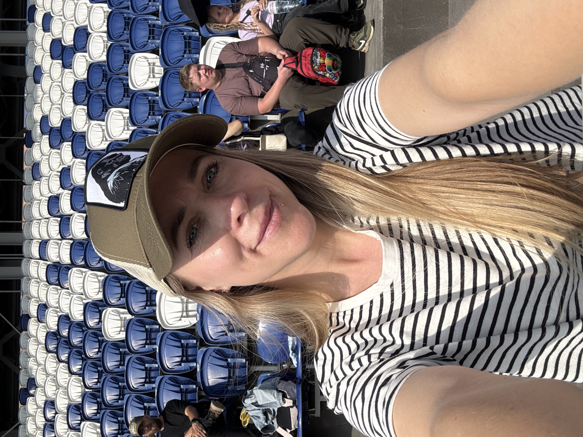 A woman in a striped shirt and baseball cap takes a selfie in a stadium with blue and white seats. A man with a beard sits to her left, and a man and a child are seated behind her to the right. The image appears to have been taken in Izhevsk, Russia, possibly at the Central Stadium.