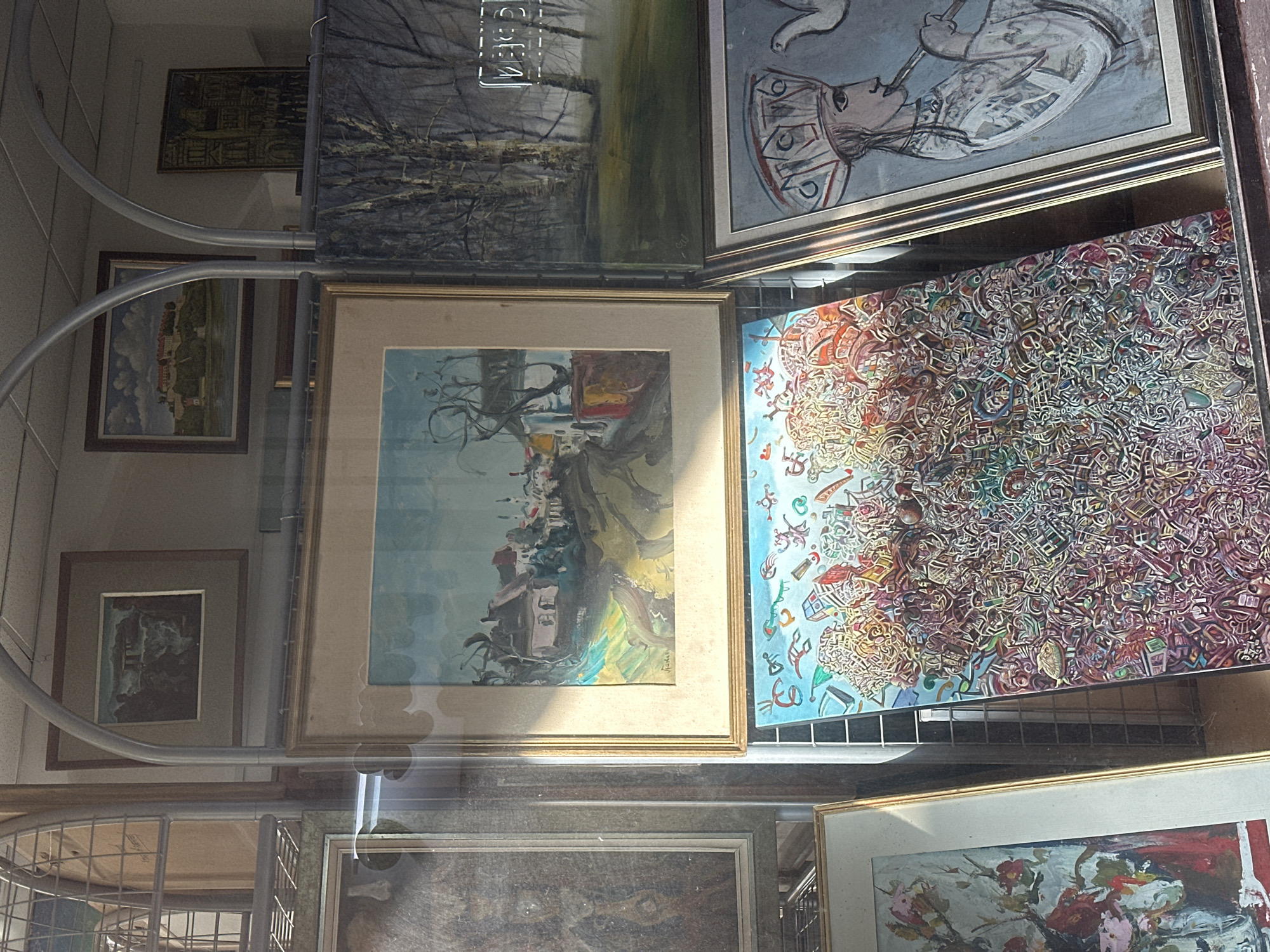 The image shows a display of various framed artworks behind glass, possibly a shop window or an exhibition in Novi Sad, Serbia. The art varies in style, from landscapes and portraiture to abstract compositions. The reflections on the glass suggest an outdoor environment, with the presence of metallic racks further obscuring a clear view of the items behind the glass.