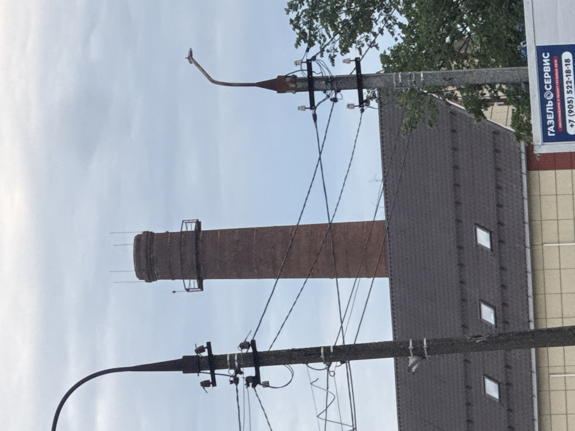 The image shows a view of a brick smokestack in the city of Dedovsk, Russia, on a cloudy day. In the foreground, a dark, curved lamppost stands to the left. To the right of the smokestack, another lamppost with a curved arm is visible. Several utility poles with electrical wires crisscross the scene. Beneath the smokestack and lampposts is the dark, sloped roof of a building with several small, square skylights. The building's facade, visible below the roofline, is light-colored with a grid pattern. A portion of a white truck or van with a blue and red sign featuring Russian text and a phone number is present in the bottom right corner. The environment appears to be urban or industrial.