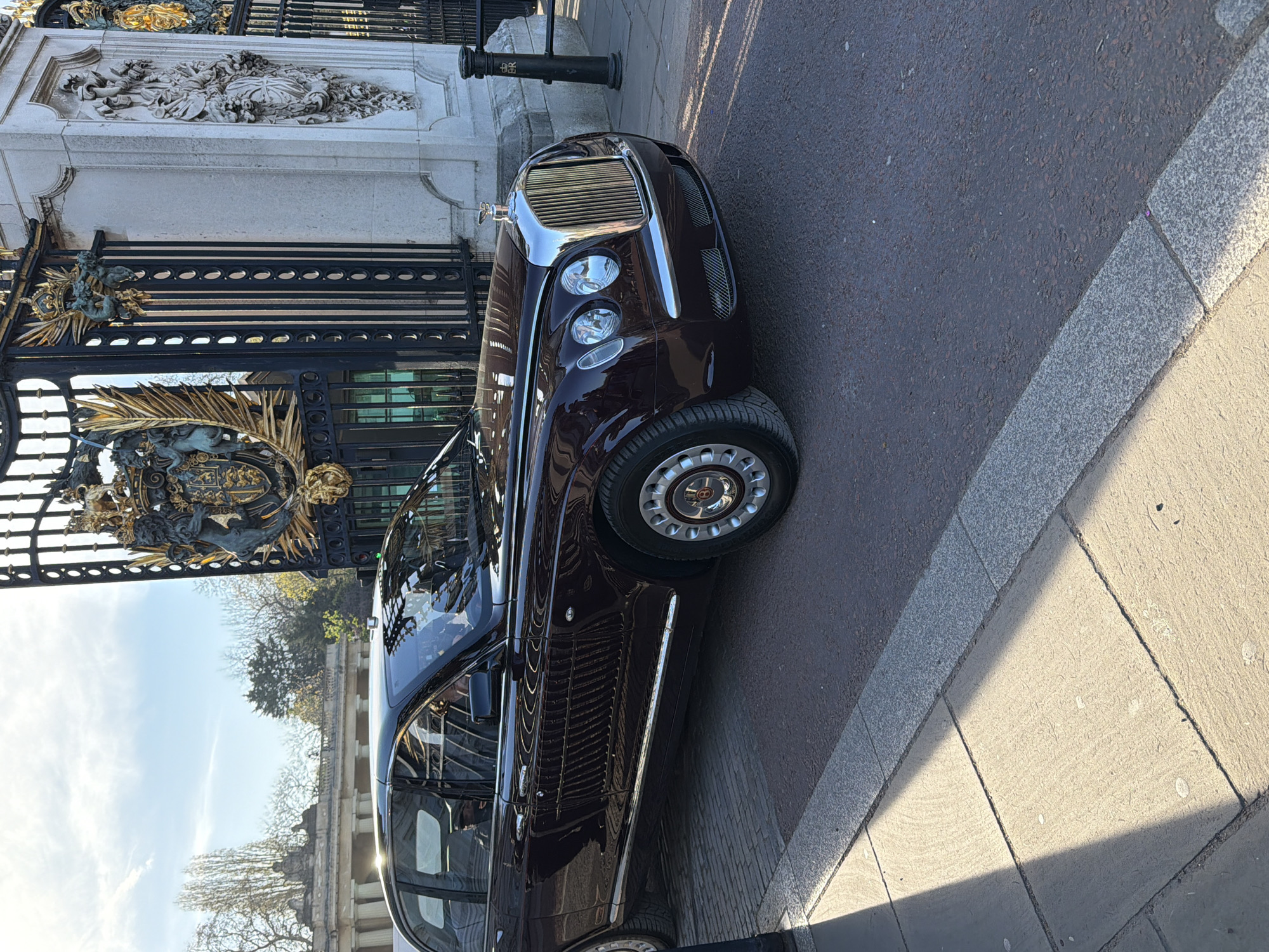 The image depicts a dark maroon Bentley limousine parked on a city street in London, adjacent to a wrought iron gate with ornate golden crests, likely indicating Buckingham Palace. The car is positioned close to the curb, with a glimpse of the palace grounds and trees visible in the background, under a partly cloudy sky. The pavement shows variations in texture and color due to sunlight and shadows.