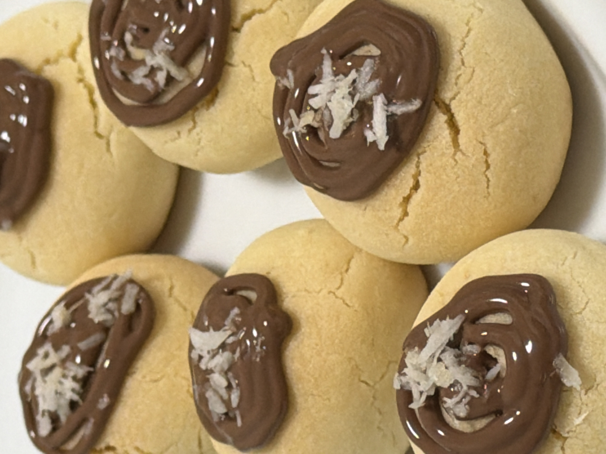 A close-up, eye-level shot shows several cookies arranged on a white surface. The cookies are light yellow and round, with a slightly cracked texture. Each cookie is topped with a swirl of melted chocolate and sprinkled with shredded coconut. The cookies are arranged in a somewhat staggered pattern, with some overlapping. The lighting is bright and direct, creating highlights on the chocolate and coconut. No people are visible in the image, and the environment is indistinct, suggesting a studio or kitchen setting. The origin of these cookies in Izhevsk, Russia, is not discernible from the visual information provided.