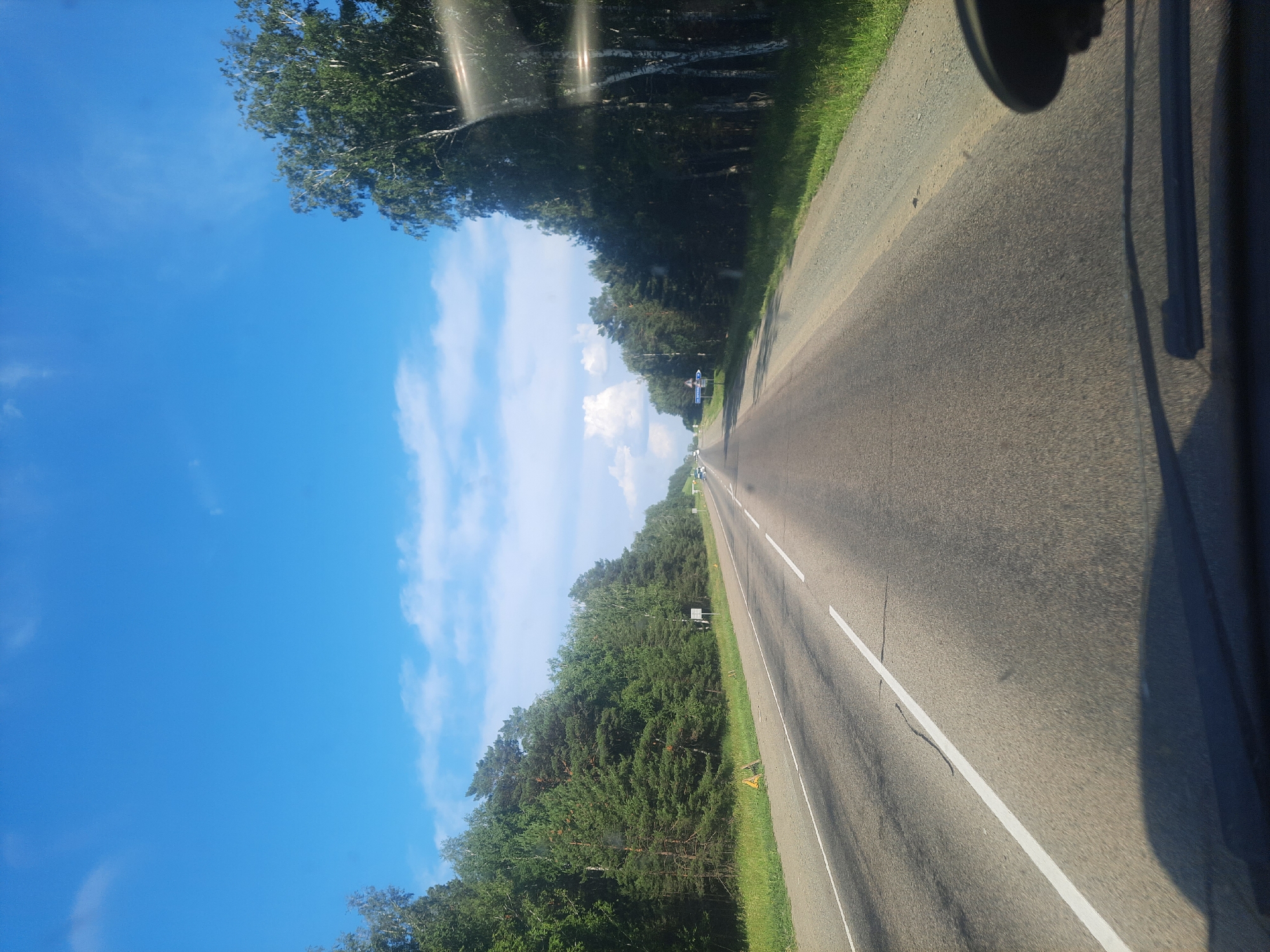 The media file captures a view from inside a vehicle traveling on a paved road through a rural landscape. The road stretches into the distance, marked by white lines. On the left side of the road, a dense forest with a variety of trees borders the asphalt. The sky is a clear blue with scattered white clouds. Sunlight creates reflections and glare on the windshield of the vehicle from which the photo is taken, casting shadows. In the distance, a few other vehicles can be seen on the road, suggesting it is in use. The time of day appears to be daytime, with bright sunlight. The overall impression is of a journey through a natural, serene environment. There is no visible text in the image. The location context provided is Баюновские Ключи, Russia.
