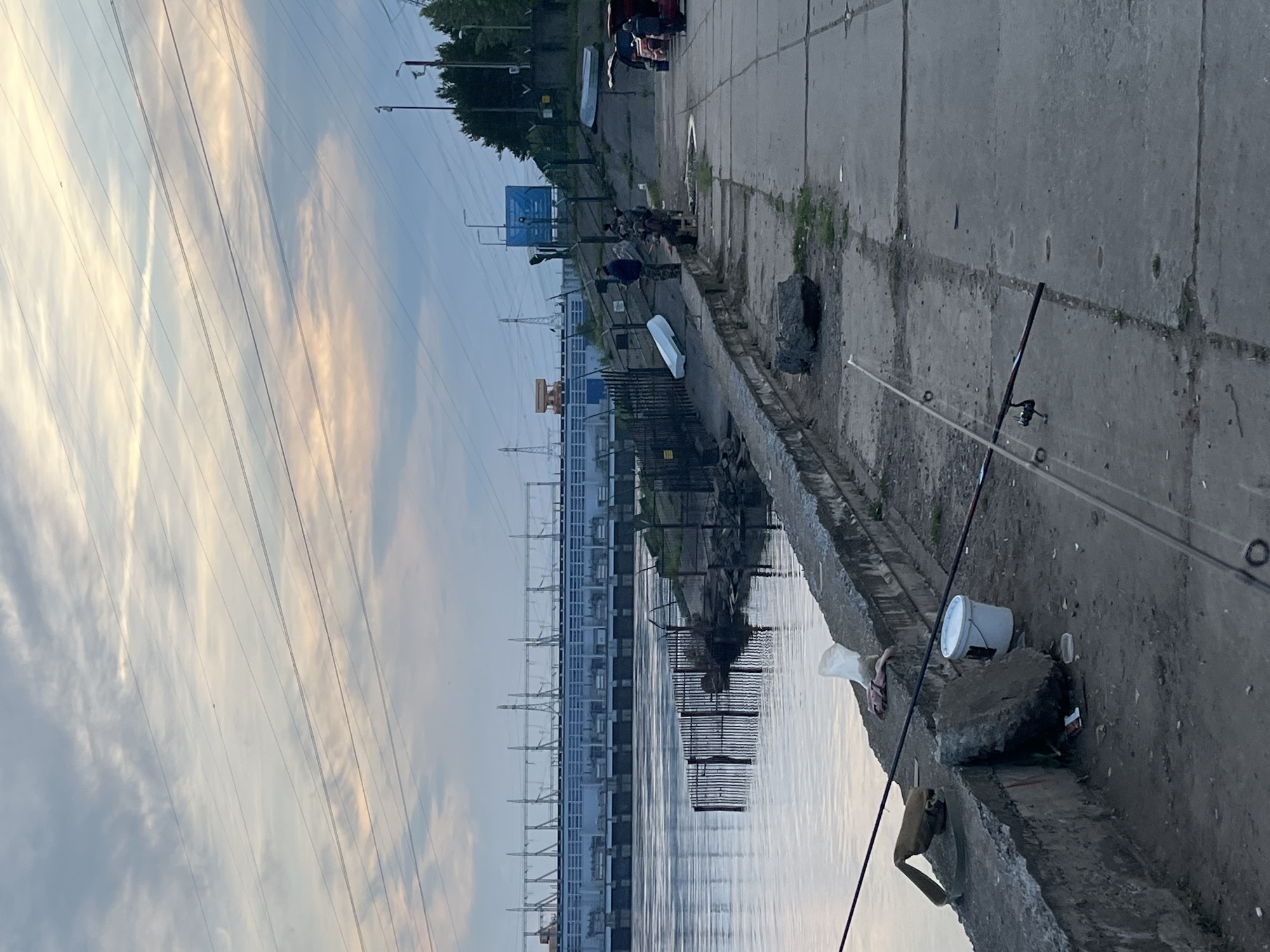 The image captures a waterside scene along a concrete embankment, likely a river or canal in Chaikovsky, Russia, during what appears to be either sunrise or sunset due to the soft, diffused light and warm hues in the sky.
In the foreground, a concrete pathway borders calm water. Several fishing rods are set up along the edge, indicating active fishing, with one prominent rod extending into the water from the lower right. A white bucket, a pair of light-colored flip-flops, and some minor litter are visible on the concrete surface near the fishing equipment.
Across the water, a large industrial complex, possibly a hydroelectric power station (consistent with the Votkinsk Hydroelectric Power Station in Chaikovsky), stretches horizontally across the mid-ground. This structure is characterized by numerous vertical elements and power line towers, with its reflection clearly visible in the still water. Further along the far bank, a paved area can be seen with a few vehicles, including a red car and a darker one, and a small white boat. A person wearing a dark top is visible standing near these vehicles. A blue sign is also present on the far bank, but its text is unreadable.
Numerous power lines crisscross the sky above the entire scene, adding to the industrial backdrop. The overall mood of the image is calm and serene, despite the presence of industrial infrastructure.