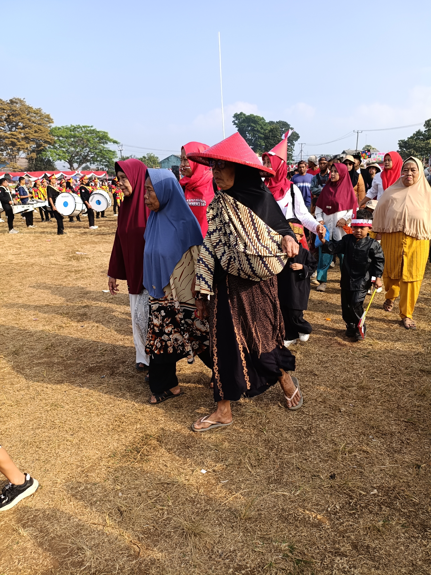 The image depicts a procession in a grassy field in Cibodas, Indonesia. Several individuals are walking, including women wearing hijabs and traditional patterned clothing, some with conical hats. A young boy in a black outfit and a red and white headband is walking next to them, holding a yellow stick. In the background, a marching band with drums is visible, along with spectators and tents. The ground is covered in dry grass, and trees are visible in the distance under a clear sky. The overall atmosphere suggests a cultural event or celebration.