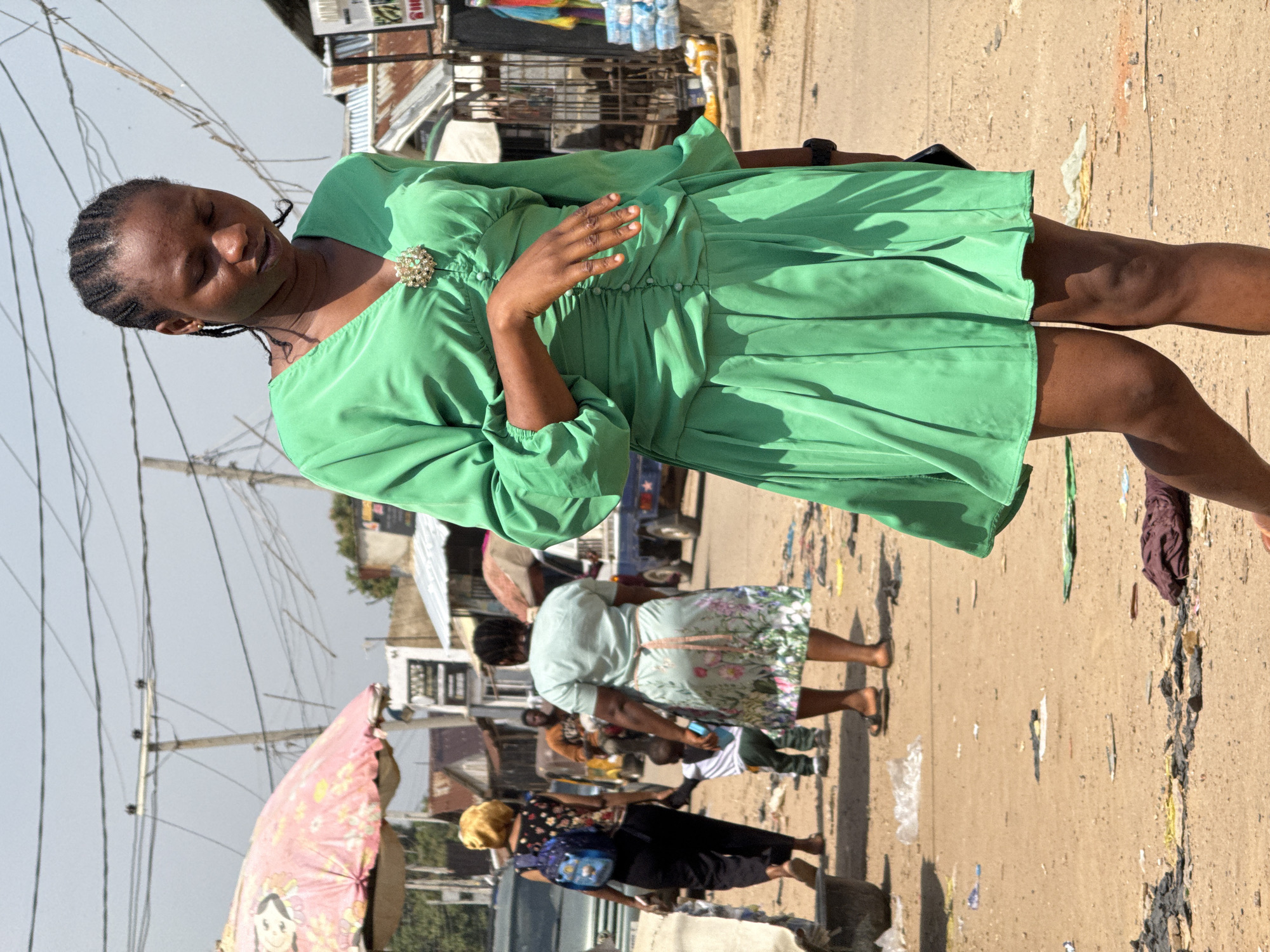 In a dusty, outdoor market scene in Abuja, Nigeria, a young Black woman in a vibrant green dress walks forward, her eyes downcast and a contemplative expression on her face. Her hair is styled in braids, and a decorative brooch adorns the neckline of her dress. The dress features a ruched bodice and a pleated skirt that ends above her knees. Her right hand rests gently on her stomach.
In the background, other people are visible, including a woman in a yellow headscarf carrying a blue backpack, a child in a striped shirt, and a woman in a floral-patterned dress. A man on a motorcycle is also present, and various market stalls and structures line the street. The ground is covered in dirt and scattered debris, with some plastic bags and fabric visible. Power lines crisscross the sky above. The scene is illuminated by bright sunlight, casting shadows on the ground.