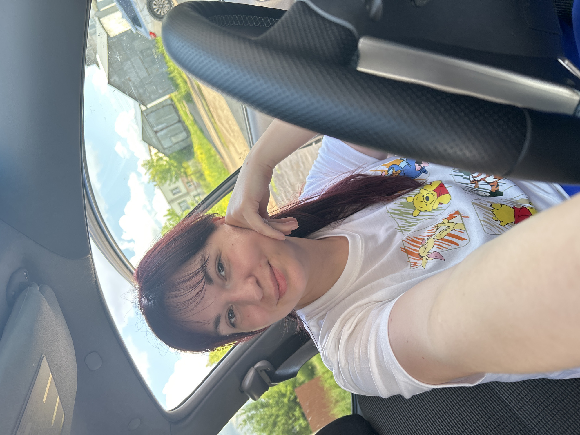 In a vehicle's interior, a woman with dark hair and bangs is pictured. She is wearing a white t-shirt with an illustration of Winnie the Pooh characters. She is resting her chin on her hand and looking at the camera. The steering wheel is visible in the foreground. Outside the car, a residential area with houses and greenery is visible under a partly cloudy sky. The location is identified as Fokinsky District, Bryansk Oblast, Russia.
