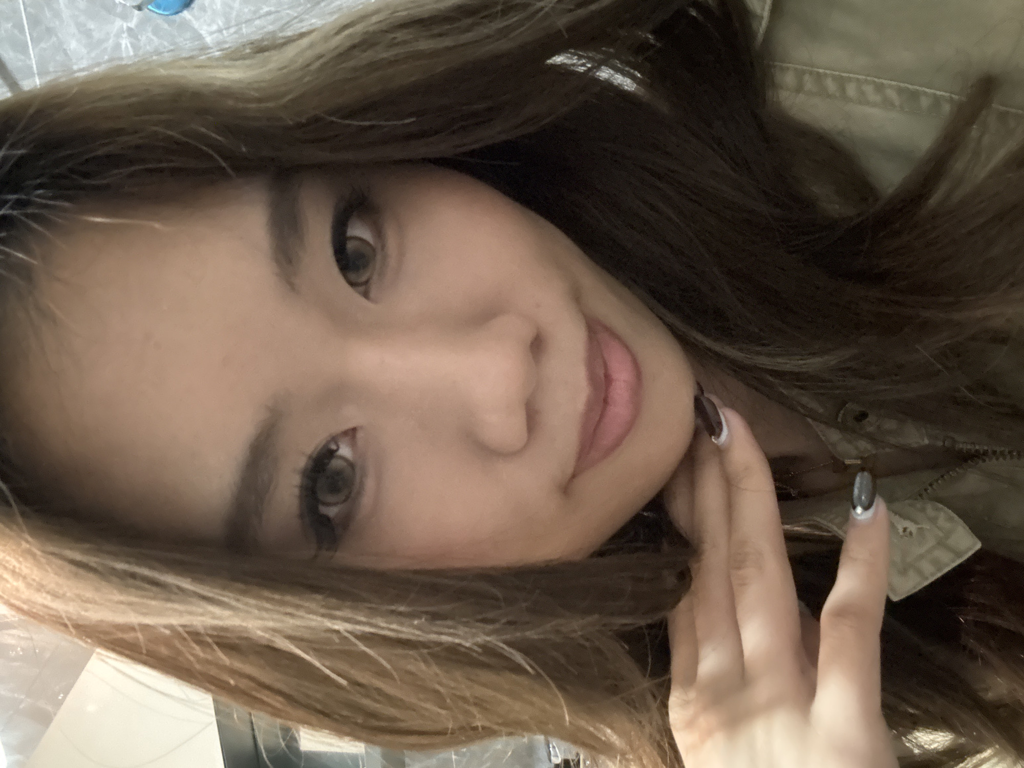 A woman with long, brown hair and light skin is holding her hand to her chin. She has brown eyes and is wearing a khaki jacket. The image is taken from a slightly low angle, and the woman is looking at the camera with a slight smile. The location is potentially Chenghua District, China, but no location-specific details are visible in the image.