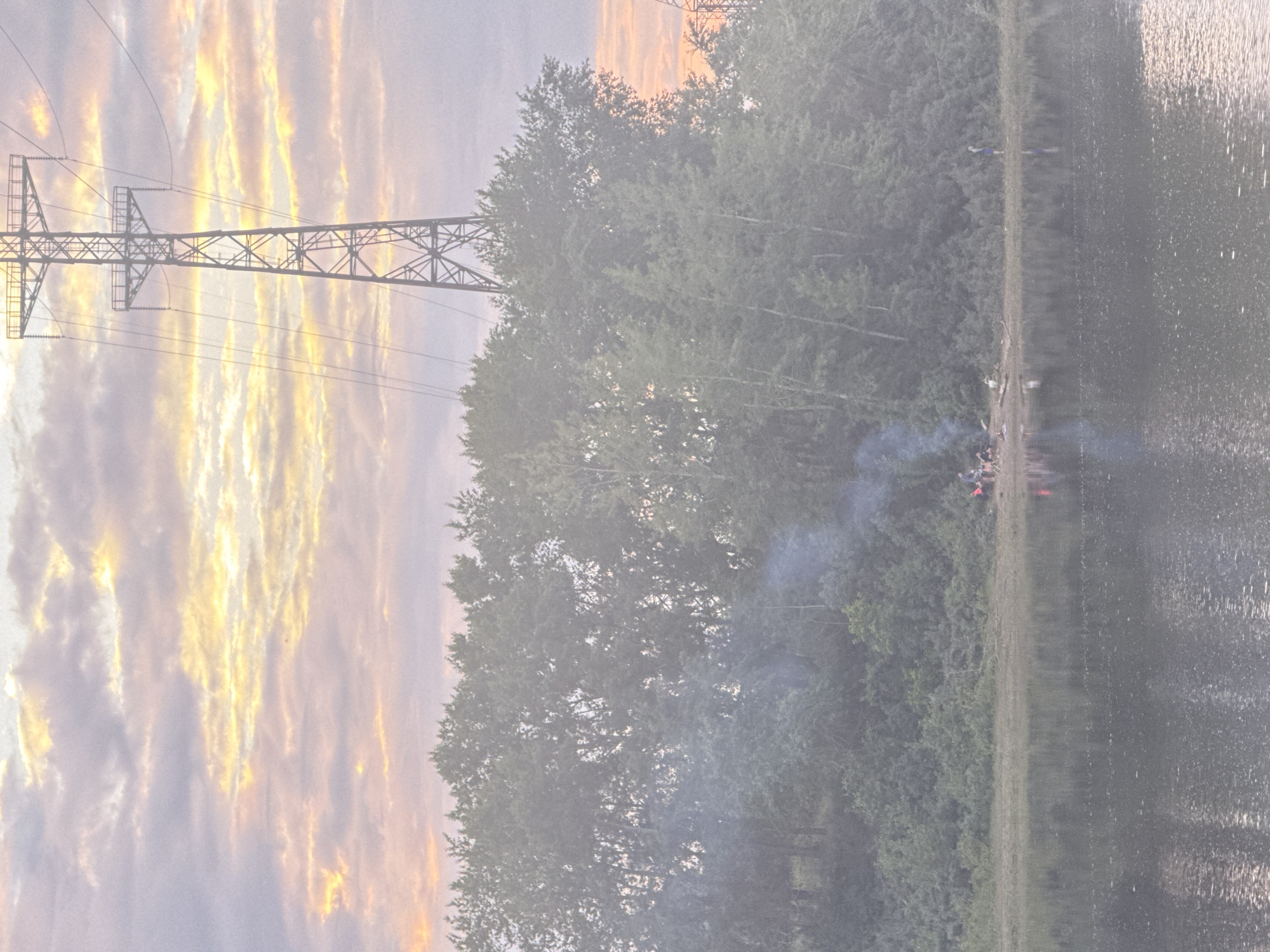 The image depicts a twilight scene on the bank of the Kama River in Chaikovsky, Russia. A tall, lattice-style electricity pylon dominates the upper right portion of the frame, silhouetted against a pale sky with wispy clouds tinged with pink and yellow hues. Dense green trees line the riverbank, with a section of the trees appearing to be slightly obscured by smoke, suggesting a campfire. In the foreground, the calm surface of the Kama River reflects the muted colors of the sky and the trees. On the riverbank, a small group of people are gathered around a fire, with some seated and others standing. To the right of the group, a single person is standing near the water's edge. The overall atmosphere is serene, with the soft light of dusk casting a gentle glow over the landscape.