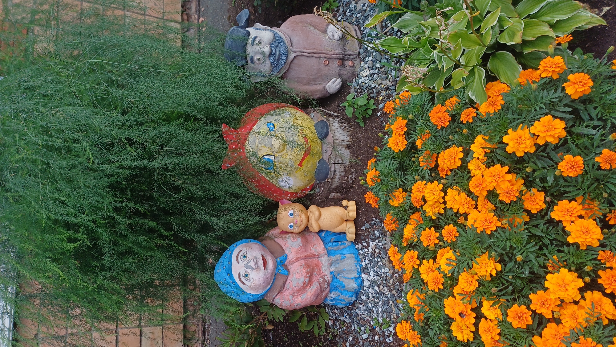 A group of garden statues are arranged in front of a dense green shrubbery. From left to right, the statues depict an elderly woman wearing a blue headscarf and a pink and blue outfit, a small, tan, cartoonish cat character with arms crossed, a round, red-hatted figure with a yellow face painted on it, and an elderly man in a brown coat and a dark cap. In front of the statues is a bed of bright orange marigolds. The ground around the statues is covered in small gray gravel. The background shows a textured, light brown brick wall. The scene is illuminated by natural daylight, casting soft shadows.