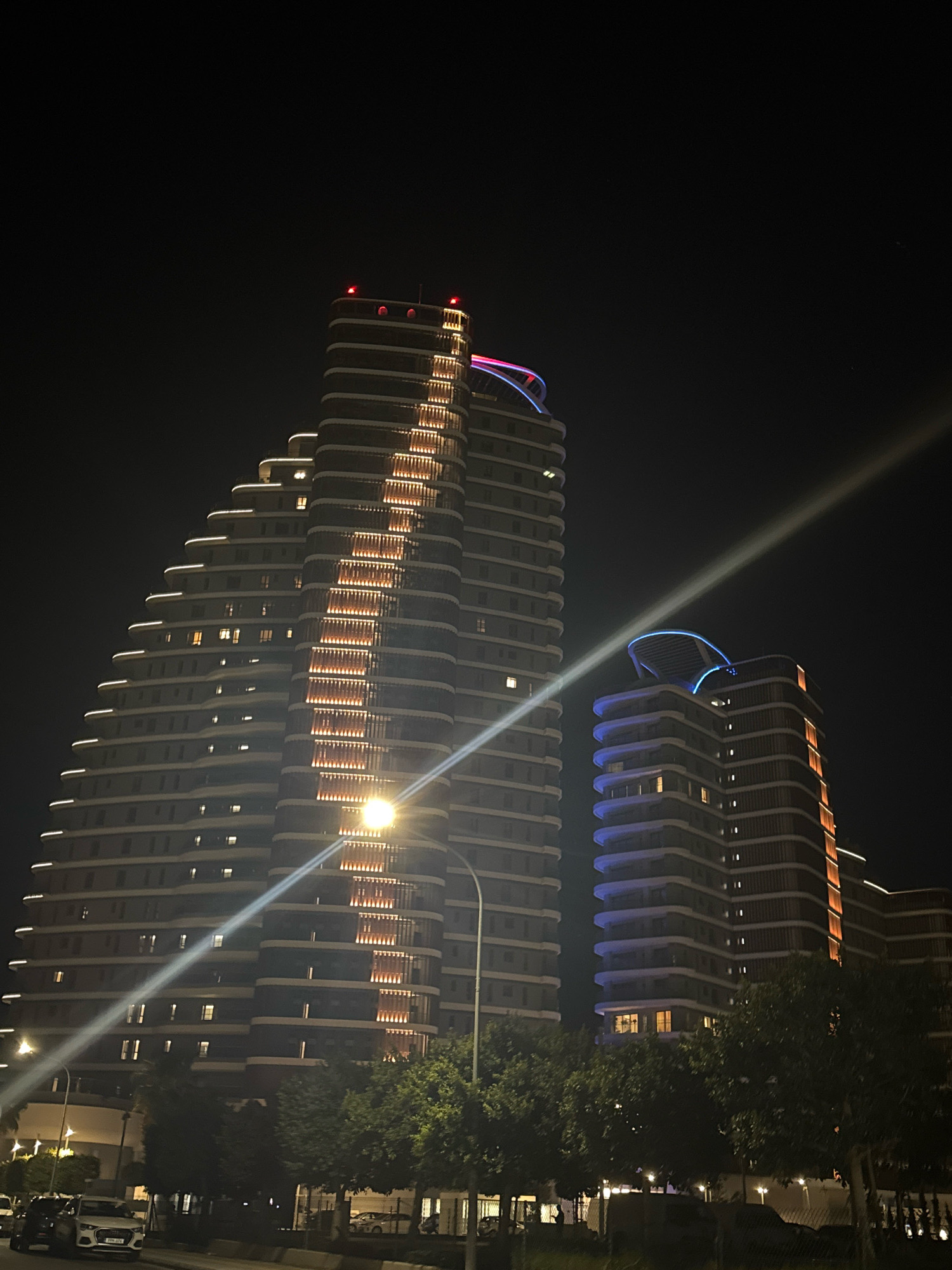 The image shows a nighttime view of two modern, multi-story buildings in Germasogeia Municipality, Cyprus. The building on the left is taller and has a tiered, stepped design with white lighting along the edges of each level. The building on the right is shorter and also features layered architecture, with blue lighting around its upper section. Both buildings are illuminated from within, with individual windows visible. A bright light beam, possibly from a spotlight or a powerful streetlamp, cuts diagonally across the frame from the lower right to the upper left. Trees with green foliage are visible in the foreground, obscuring the lower portions of the buildings and the ground. A street and parked cars are faintly visible at the bottom of the image. The overall scene depicts an urban night landscape with distinctive architectural lighting.