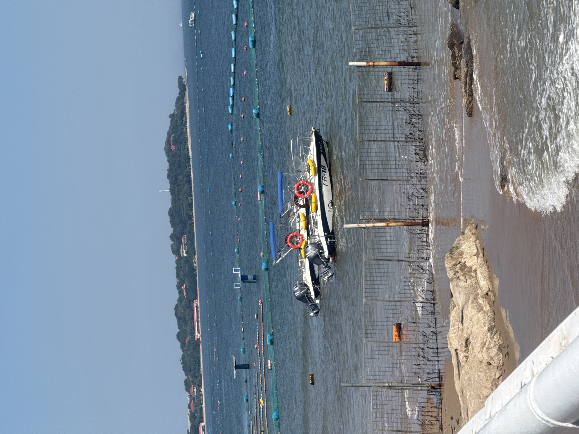 The image depicts a sunny day at a coastal area in Qinhuangdao, China. The foreground shows a rocky shoreline with waves lapping at the sand, bordered by a metal fence. In the middle ground, two white motorboats with outboard engines are moored in the blue water. One boat is closer to the viewer and has a red life preserver and yellow buoys visible. The boat in the distance is partially obscured. The background reveals a lush green island with trees and buildings, stretching across the horizon under a clear blue sky. Numerous blue and red buoys, connected by netting, dot the water, suggesting aquaculture or a marine facility.
