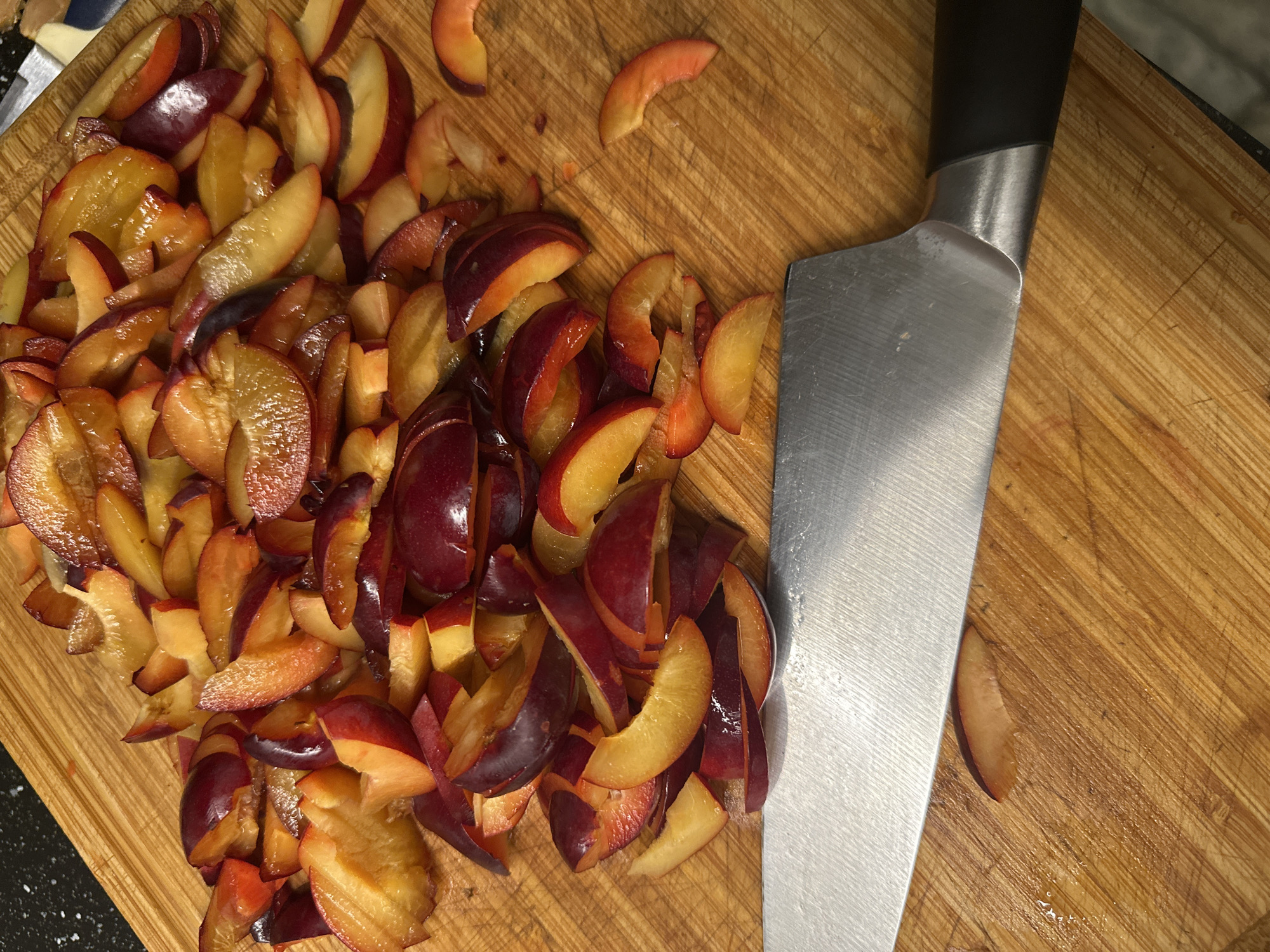 A close-up, overhead shot shows sliced plums on a wooden cutting board. A chef's knife rests in the foreground, its blade pointing towards the right. The plums, varying in color from deep purple to bright yellow and orange, are scattered across the board, some whole, some in halves, and many in wedges. The cutting board exhibits visible grain and scratch marks from previous use. The lighting suggests an indoor setting, likely a kitchen. The image does not contain any elements that can be used to determine its location in Nevinnomyssk, Russia.