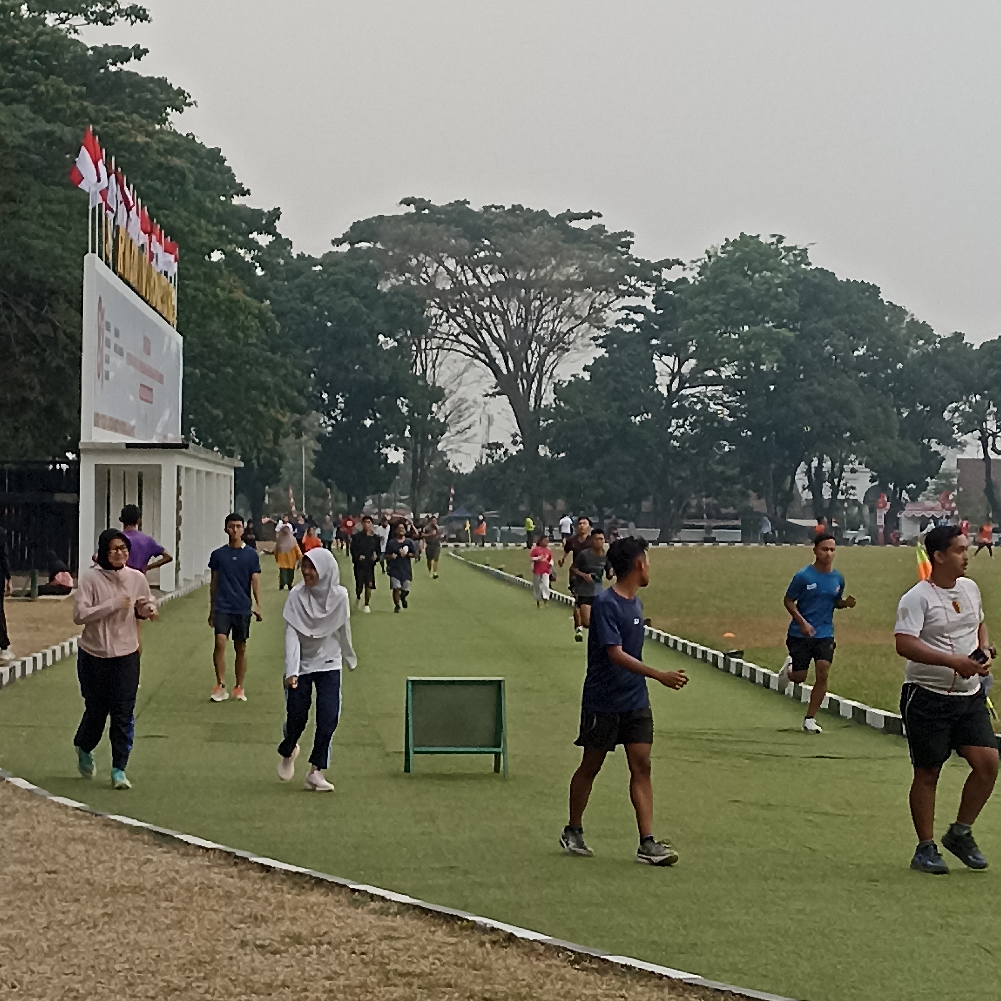 This is a medium-shot, eye-level, outdoor photograph taken in the city of Magelang, Indonesia, likely at a sports field or track. The scene depicts a group of people engaged in physical activity, primarily running and walking, on a bright green artificial turf track. The background is dominated by lush green trees and a light gray, overcast sky.
In the foreground, several individuals are visible. On the left, a woman in a pink jacket and dark pants is jogging, her body angled slightly towards the viewer. Next to her, a woman in a white hijab and white long-sleeved top is also jogging, looking ahead. A man in a blue t-shirt and black shorts is running in the middle ground, his gaze forward. To the right, a man in a white t-shirt and black shorts is walking, holding what appears to be a phone in his right hand. Further back, a larger group of people are scattered across the track, running and walking in various directions. Some are wearing athletic attire, while others are in casual clothing.
To the left of the image, a white structure with a large screen or banner is visible, adorned with Indonesian flags. The banner has text and graphics, but they are not clearly discernible. The track is bordered by a low white and black barrier. In the background, a green wooden stand is positioned on the track. The overall atmosphere suggests a public event or a casual exercise session.