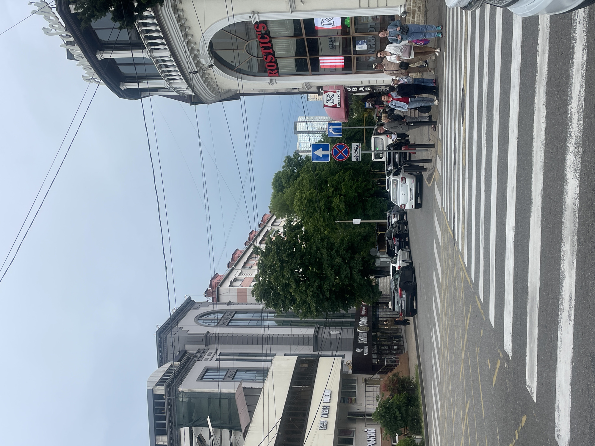 This image depicts a street scene in Sochi, Russia. A crosswalk with pedestrians crosses the street. A building with the name "ROSTIC'S" is visible at the upper edge. There are various traffic signs indicating rules of the road. Cars are parked along the side of the street. Trees and other buildings line the street.