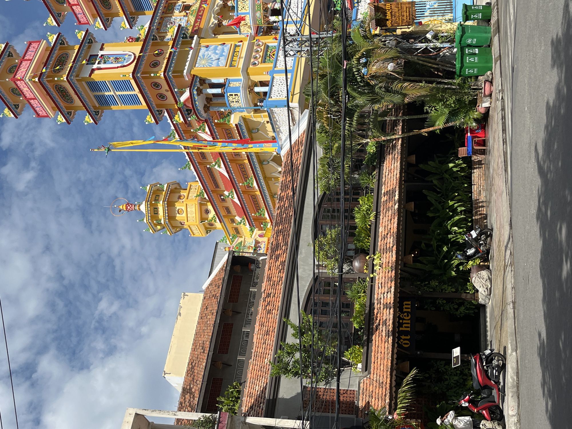 This is a photograph of the Tinh Do Lam Son temple, a prominent Buddhist temple located in Nha Trang, Vietnam. The temple is characterized by its vibrant yellow and ornate architectural design, featuring multiple tiered towers adorned with colorful decorations and Buddhist imagery. In the foreground, a street scene unfolds. A red scooter is parked on the sidewalk to the left of the frame, and a person in a white garment stands beside it. To the right, several green trash bins are lined up along the sidewalk. Behind the street level, a building with a terracotta tiled roof and lush greenery on its balconies suggests a commercial establishment, possibly a restaurant, with outdoor seating visible. Palm trees and other tropical plants are scattered throughout the scene, adding to the tropical ambiance. The sky above is a clear blue with scattered white clouds. The overall impression is a blend of religious architecture and daily urban life in a tropical city.