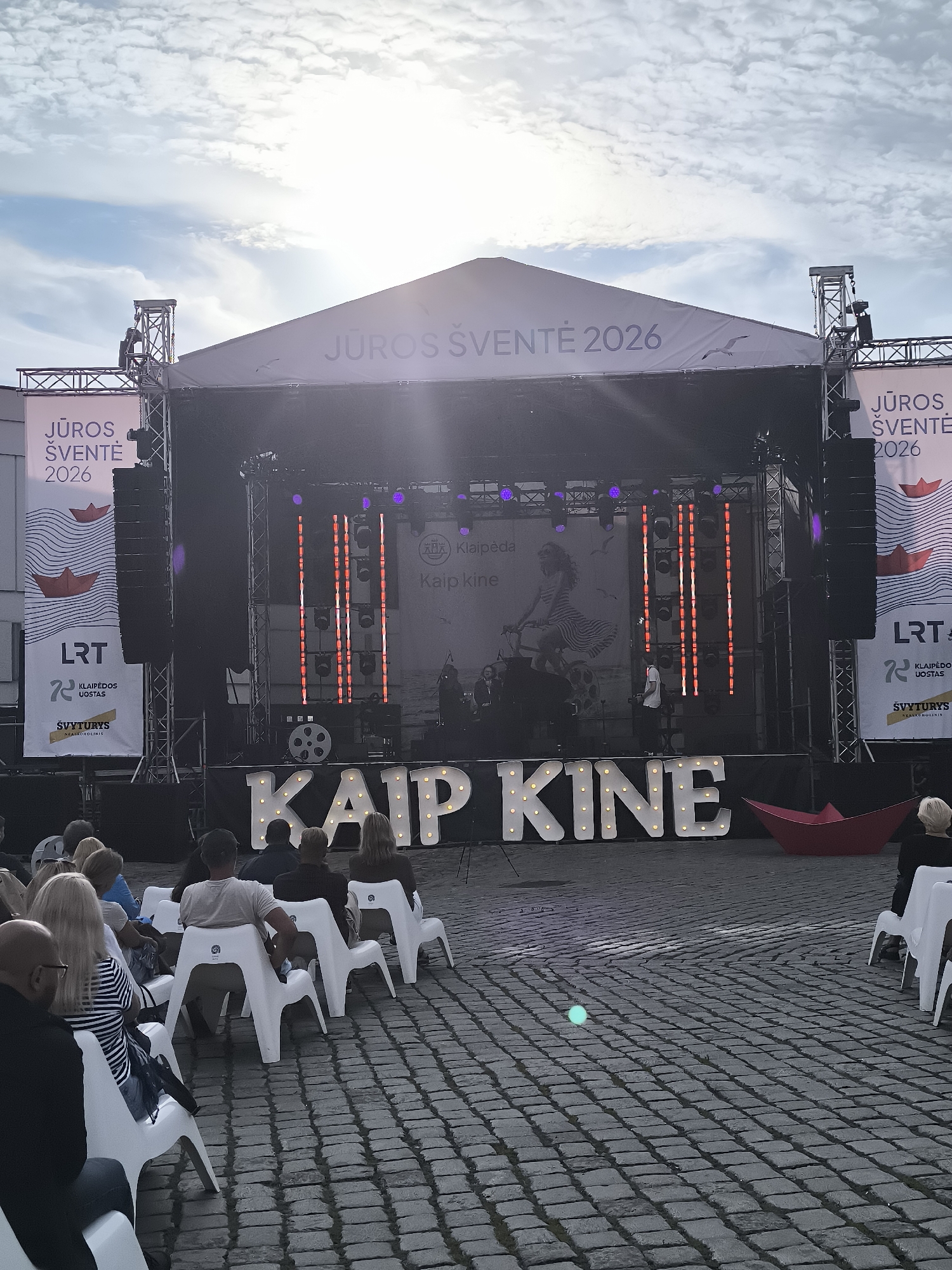 The image depicts an outdoor event, likely a festival or concert, taking place in Klaipėda, Lithuania, specifically at the Klaipėda Seaport, as indicated by signage. A large stage is set up with a prominent banner at the top displaying "JŪROS ŠVENTĖ 2026," which translates to "Sea Festival 2026." The stage features lighting rigs, speakers, and a large illuminated sign spelling out "KAIP KINE," possibly a part of the event's theme or name. On the stage, there are musicians or performers, though they are largely obscured by the lighting and the distance.
In front of the stage, an audience is seated on white plastic chairs arranged on a cobblestone plaza. The audience members are facing the stage, with some visible from behind. The scene is lit by the stage lighting and the natural light from the sky, which is partly cloudy. The overall atmosphere suggests a public gathering for entertainment.
Additional signage on the sides of the stage includes logos for "LRT," "KLAIPĖDOS UOSTAS" (Klaipėda Seaport), and "ŠVYTURYS," a Lithuanian brewery. A red paper boat decoration is visible near the stage, echoing the nautical theme. A small glowing orb is present on the cobblestones in the foreground.