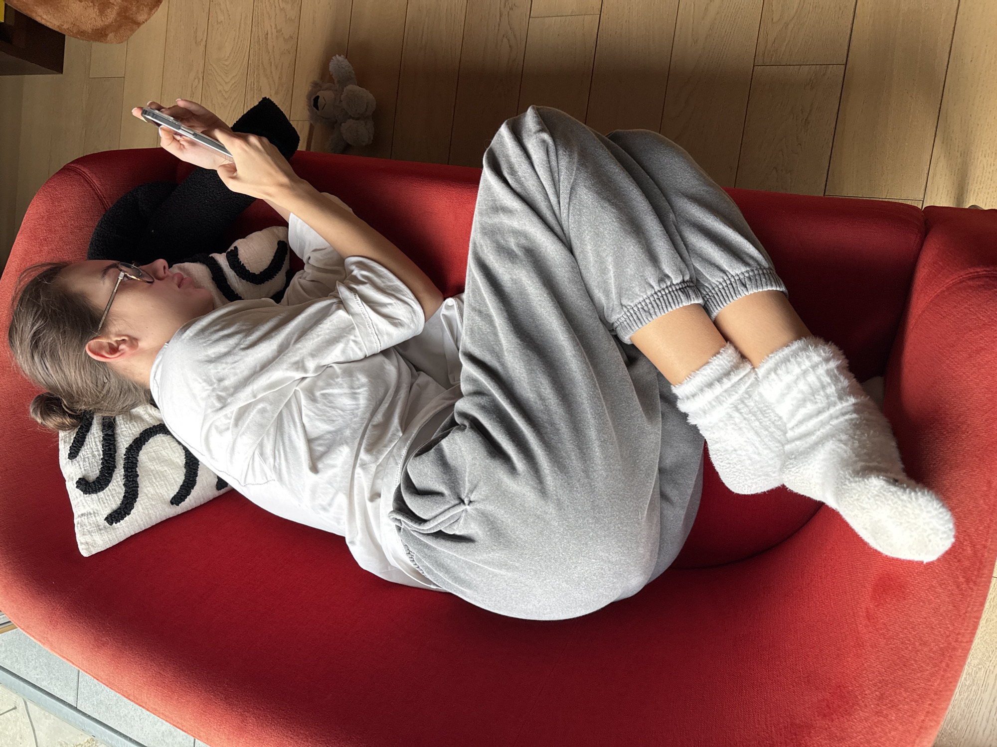 A young woman lies diagonally on a red couch with wooden flooring visible in the background, suggesting an indoor setting. She is dressed in a white t-shirt, grey sweatpants, and white socks. She's holding a smartphone in both hands and appears to be looking at it. There is a white pillow with black patterns behind her head, and a black cushion next to the pillow. A small stuffed animal is visible near the couch. The photograph was taken from above. No specific details confirm it is Warsaw, Poland.