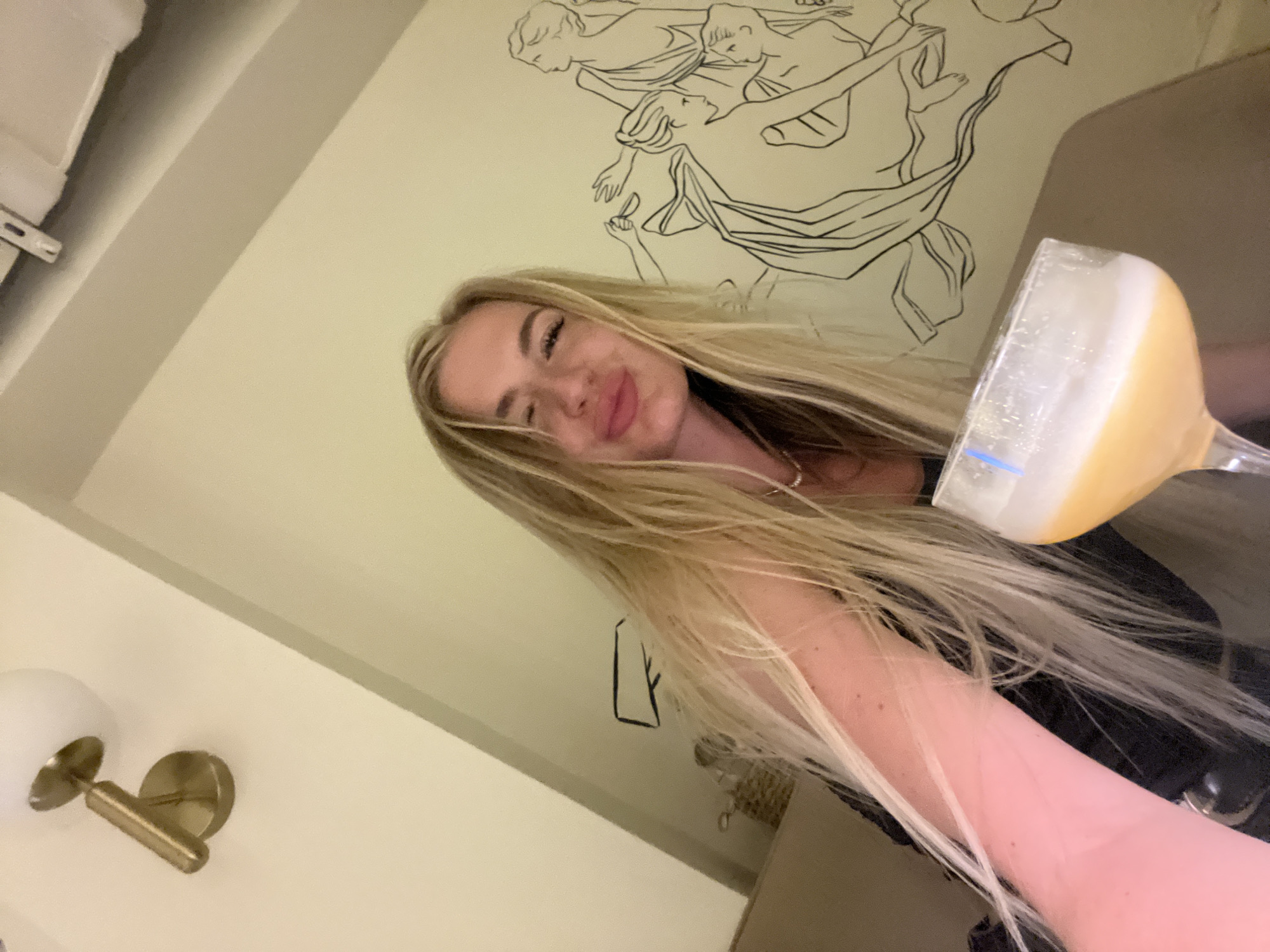 A blonde woman is holding a glass of a yellow beverage in front of a wall with an illustration of nude figures. The photograph was likely taken indoors. A light fixture is visible in the lower left corner of the frame. The location is Boreti, Montenegro.