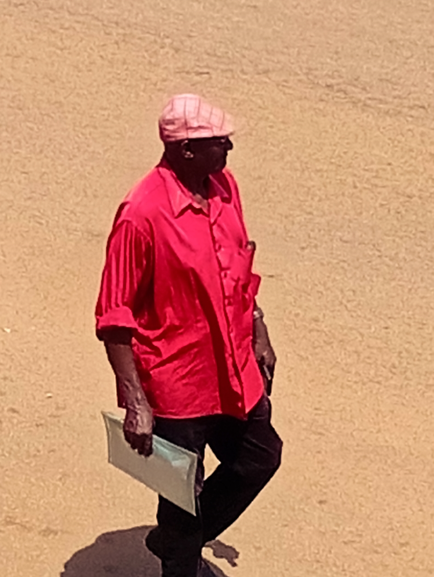In the city of Bukuru, Nigeria, a Black man is walking across a sandy, outdoor area. He is wearing a pink flat cap and a bright red collared shirt with the sleeves rolled up. He is holding a light-colored folder or envelope in his left hand. He is also wearing black pants. His shadow is visible on the ground.