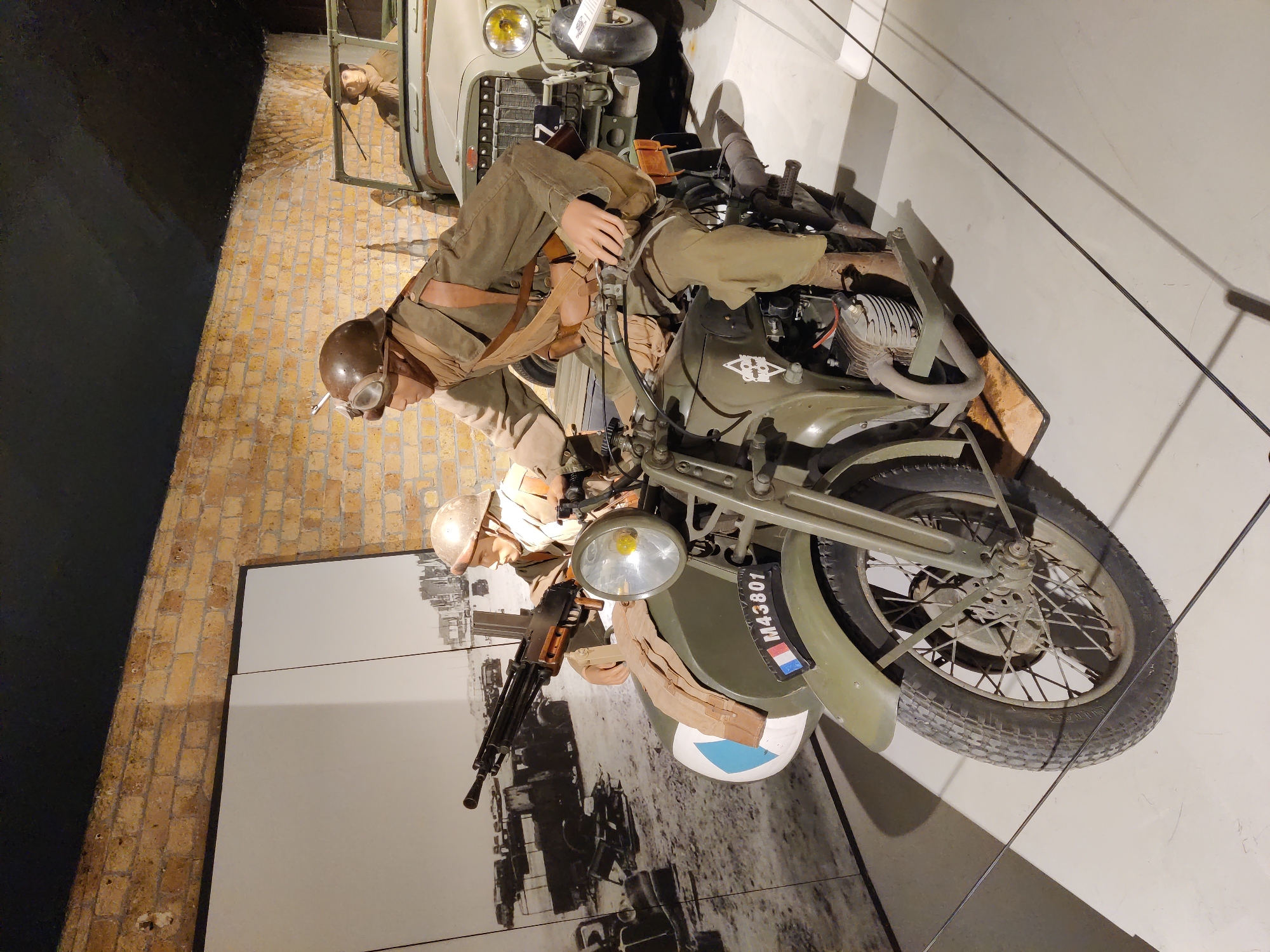 The image displays a museum exhibit with mannequins dressed as soldiers on a motorcycle with a sidecar and a jeep in the background. The motorcycle and sidecar are olive green, with the sidecar featuring a French flag and the license plate "M43801". A mannequin in a military uniform, helmet, and goggles is seated on the motorcycle, holding a machine gun. Another mannequin is visible in the sidecar, and a third mannequin is in the jeep behind them. The exhibit is set against a backdrop of a large black and white photograph depicting a war scene with damaged vehicles and buildings. The background wall is made of brick. The overall scene is designed to represent a historical military setting, possibly alluding to events in Dunkirk, France, given the presence of French military elements and the context of wartime imagery.