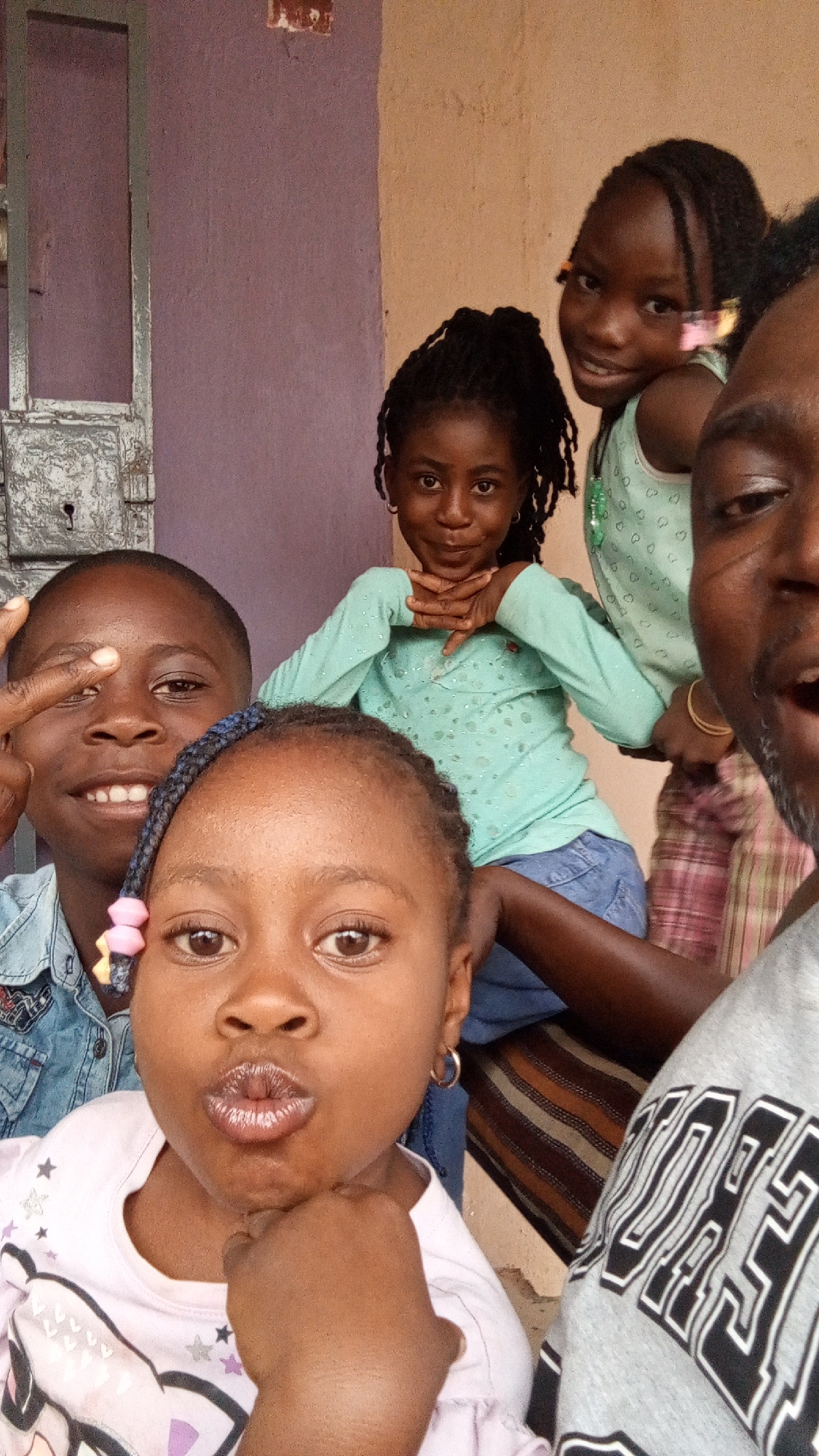 A group of five people, likely a family or friends, are captured in a selfie-style photograph taken outdoors.
Foreground: A young girl with braided hair decorated with a pink and white flower holds her chin and puckers her lips towards the camera.
Middle Ground: Behind her, another young boy in a denim shirt smiles with his fingers raised. To the right and slightly behind him, a girl wearing a turquoise long-sleeve shirt holds her chin in her hands. A third girl wearing a similar green shirt and standing behind the seated girl smiles at the camera.
Background: On the right side of the image is a man in a gray shirt. Behind the group is a building with a lavender-colored wall and a silver door. The picture was taken in Sabon Tasha, Nigeria.