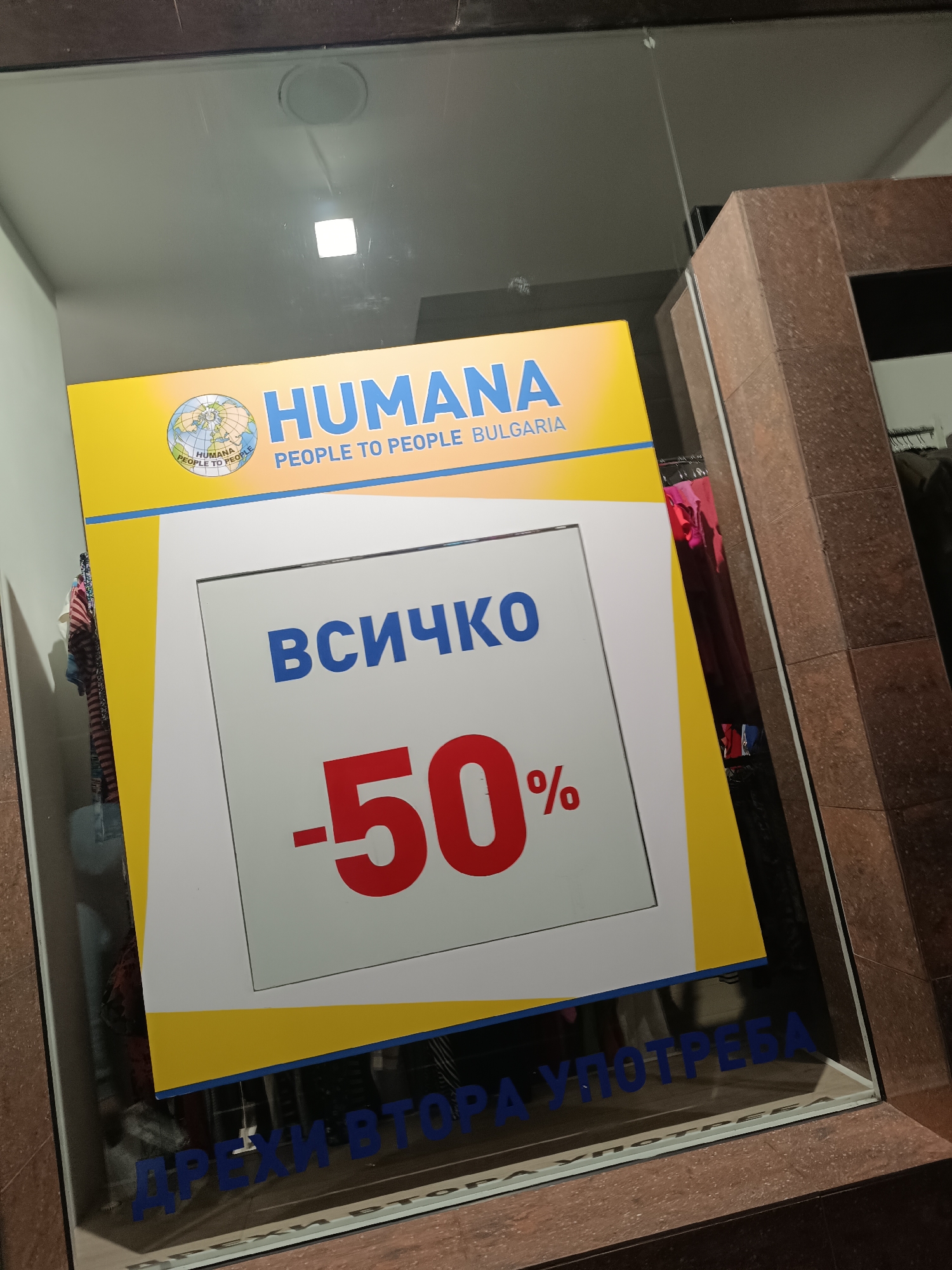 A large yellow and white promotional sign for a "Humana" store is prominently displayed in a storefront window, likely in Pleven, Bulgaria. The sign is angled, partially reflecting the interior ceiling with a square light fixture. The "Humana" logo features a globe with "HUMANA PEOPLE TO PEOPLE" written around it, and the full name "HUMANA PEOPLE TO PEOPLE BULGARIA" is visible. The main message on the sign, written in bold blue Cyrillic text, states "ВСИЧКО" (VSIČKO), meaning "ALL" or "EVERYTHING," followed by a large red "-50%," indicating a fifty percent discount on all items. Below this, partially obscured by the bottom frame of the sign, faint blue Cyrillic text on the glass reads "ДРЕХИ ВТОРА УПОТРЕБА" (DREHI VTORA UPOTREBA), meaning "SECOND-HAND CLOTHES." Through the clear glass, behind the sign, clothes on racks are visible inside the store, suggesting it is a second-hand clothing retail establishment. The setting appears to be an urban commercial environment.