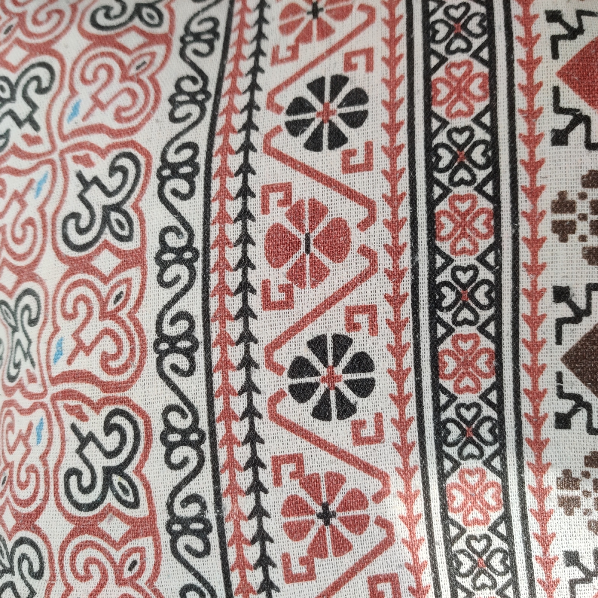 This is a close-up, top-down shot of a patterned fabric. The fabric is white with a repeating design in red, black, and a few small blue accents. The pattern features decorative scrollwork at the top, followed by a row of pointed, arrow-like shapes in red and black. Below this is a band with geometric designs and floral motifs in red and black. The bottom section shows another row of the arrow-like shapes and a more complex geometric pattern with brown and black elements. The image focuses solely on the texture and details of the fabric. There is no identifiable location within Yekaterinburg, Russia, as the image contains only fabric.