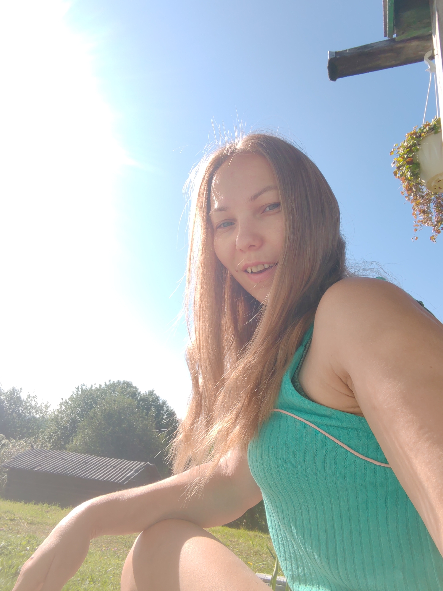 The image shows a young woman with long, light brown hair and blue eyes. She is wearing a turquoise, ribbed tank top. The woman is outdoors, with a bright blue sky and bright sunlight in the background. To her left, there is a wooden structure with a dark, corrugated roof and green trees behind it. To her right, a hanging planter with green and reddish-brown foliage is attached to the side of a building. The overall setting suggests a rural or suburban environment, possibly a backyard or porch area. There is no specific indication that the location is in the city of Чухлэм, Russia; it is a general outdoor scene.