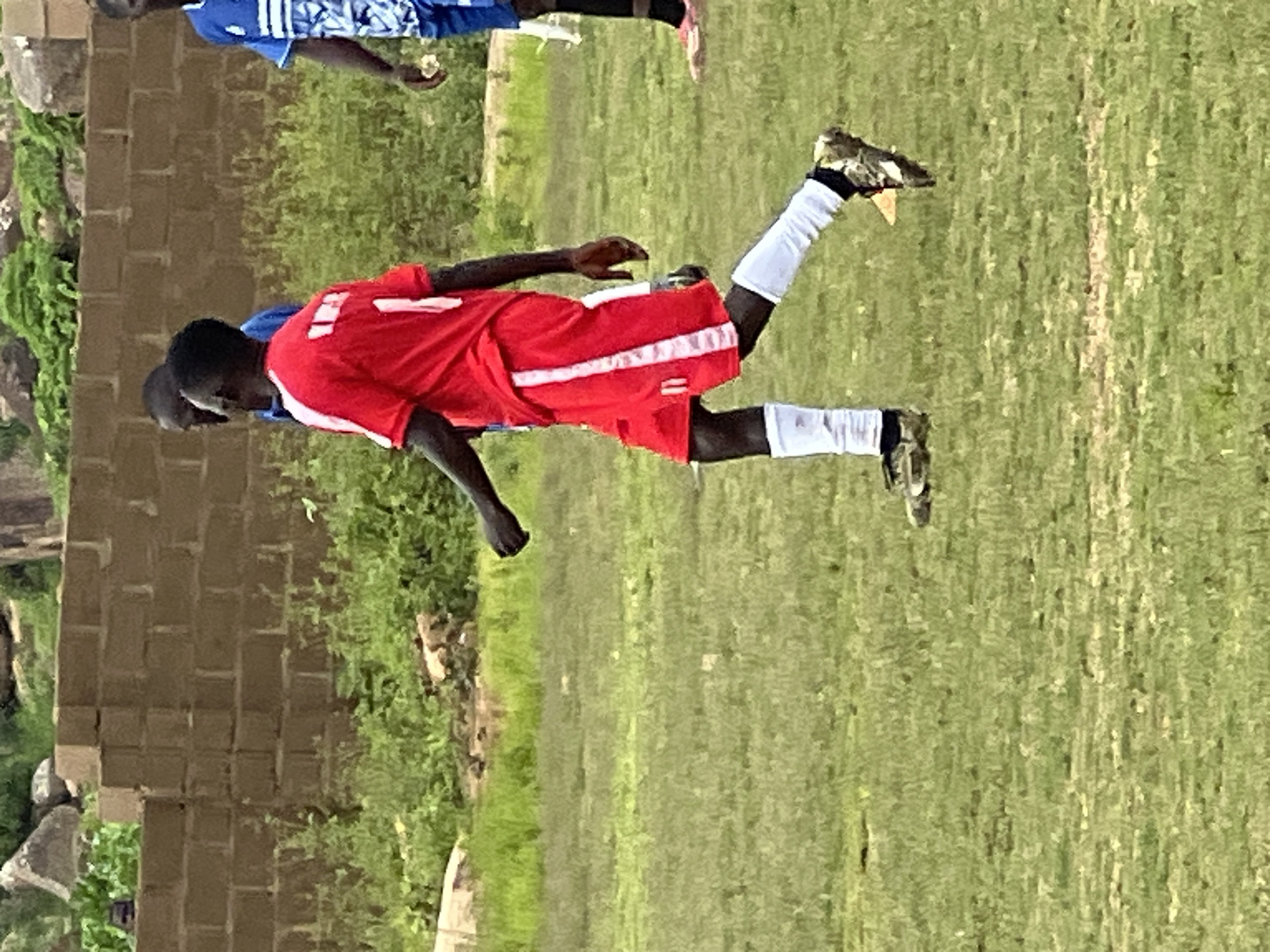 A young Black male soccer player, wearing a red jersey with white trim and the number 11 on the shorts, is captured in motion on a grass field. He is wearing white socks and patterned soccer cleats. To his left, another player in a blue jersey is partially visible. In the background, a low brick wall and lush green vegetation suggest an outdoor setting. The overall scene conveys a moment from a soccer match or practice. The image appears to have been taken in Jos, Nigeria.
