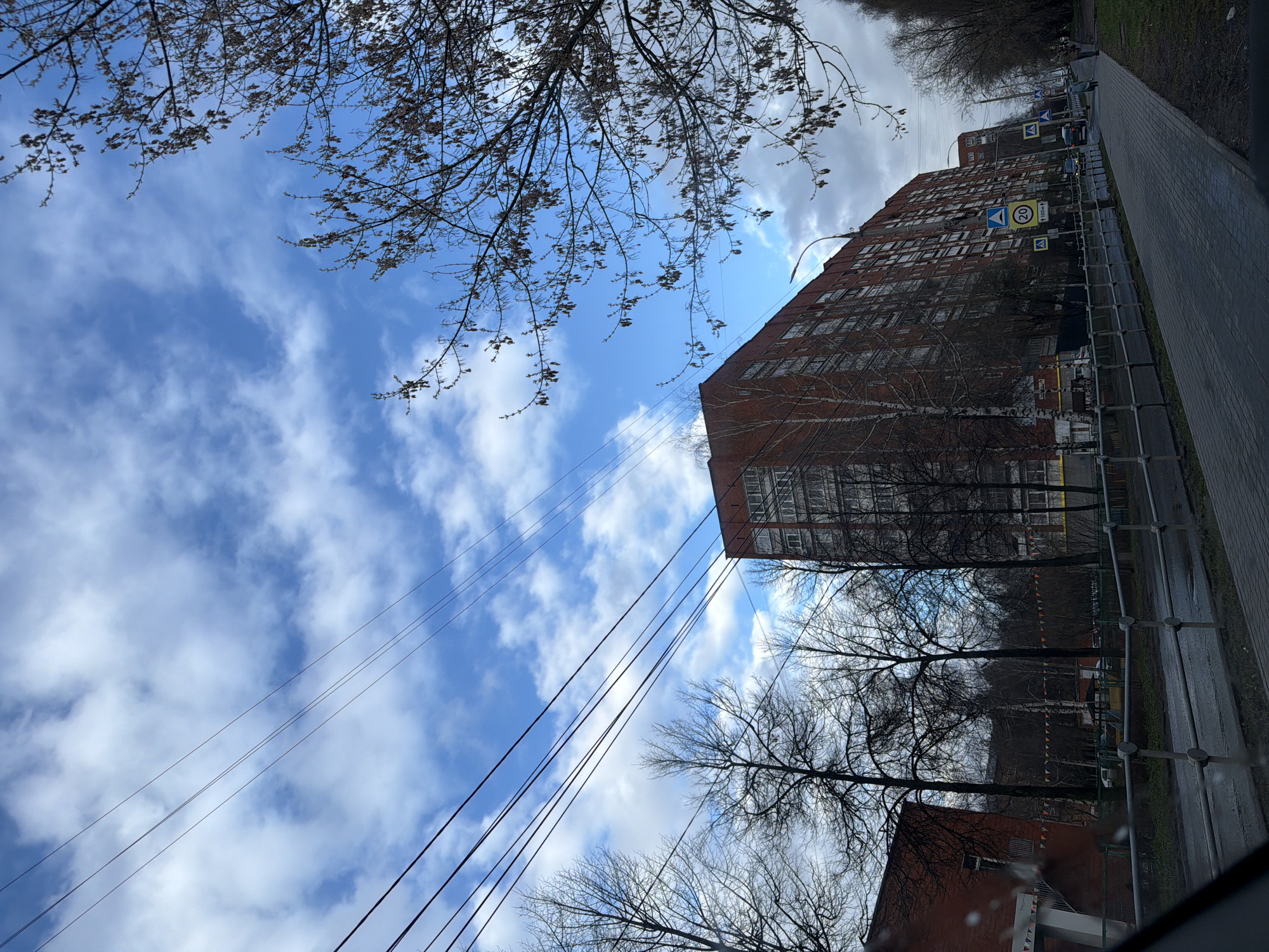 The image displays an urban street scene in Izhevsk, Russia, captured during daylight hours under a bright sky with scattered white clouds. The perspective looks upwards, partially framed by bare tree branches in the upper left and center foreground, suggesting late autumn, winter, or early spring. Multiple black utility wires run diagonally across the sky from left to right.
A large, multi-story building, constructed of reddish-brown brick with numerous rectangular windows, dominates the right side of the frame. Adjacent to the building, a paved street or sidewalk stretches into the distance, lined with a low metal barrier. Several vehicles are visible on the street, and faint figures that appear to be people can be seen walking in the distance along the sidewalk.
Prominent road signs include a blue circular sign with a white border displaying "20," indicating a speed limit of 20 km/h. Below it, a smaller white rectangular sign with an arrow and "200m" is visible, likely indicating distance. Further down the street, two yellow square signs with a black walking person symbol denote pedestrian crossings. Bare trees also line the street further into the background, with another building partially visible.