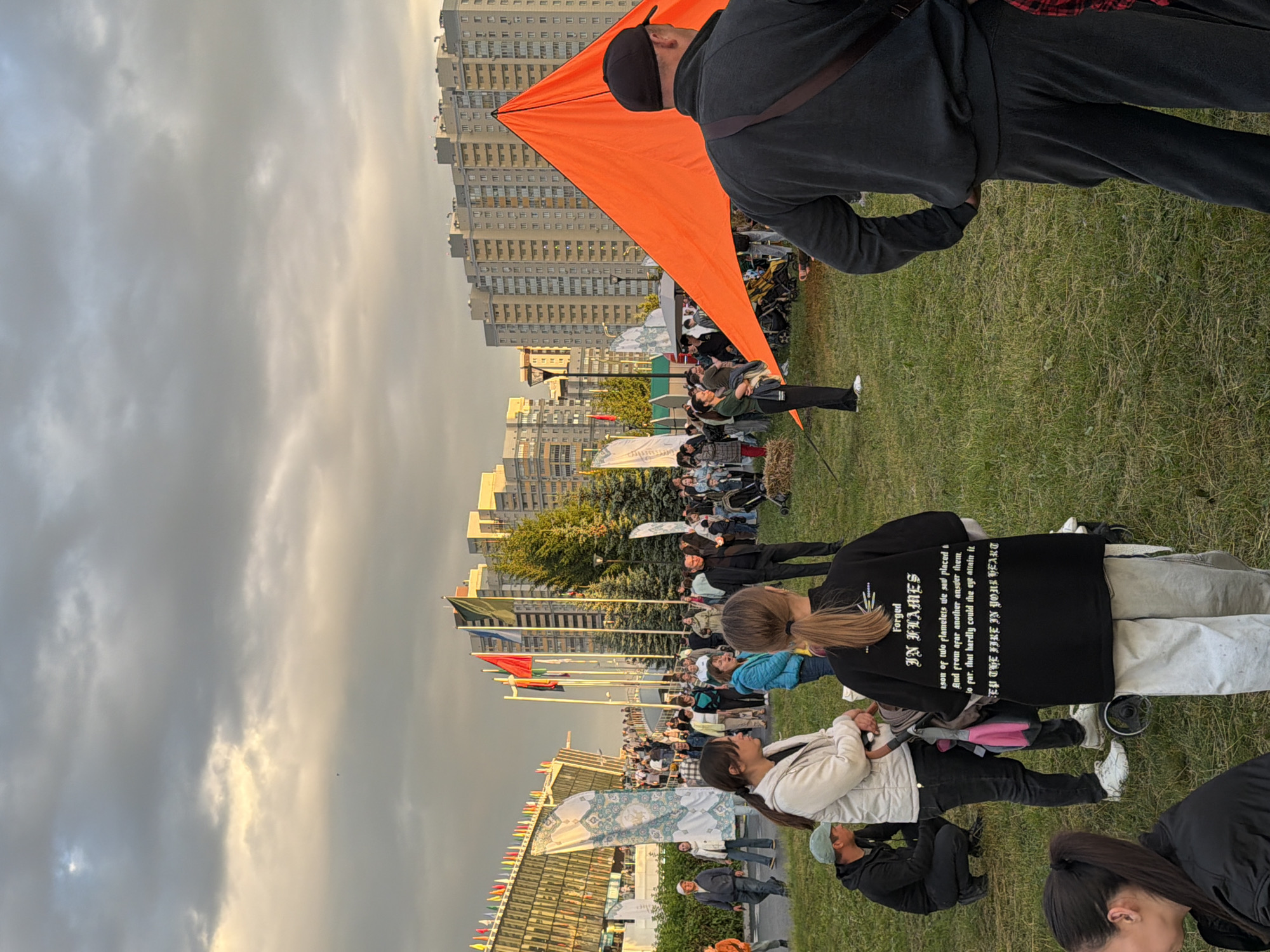 This is an outdoor scene in the city of Unknown, Unknown, with a cloudy sky and late afternoon lighting. A crowd of people are gathered on a grassy area in front of a modern building complex and tall trees. A large, bright orange, tent-like structure is prominently displayed. Several flags are visible, including a line of colorful flags along the edge of the modern building. People are standing, walking, and some are seated. One person in the foreground is wearing a black t-shirt with white text that reads "Forged IN FLAMES" and further text below. Another person in the foreground is facing away from the camera, wearing a black jacket and black pants. The overall atmosphere appears to be a public gathering or festival.