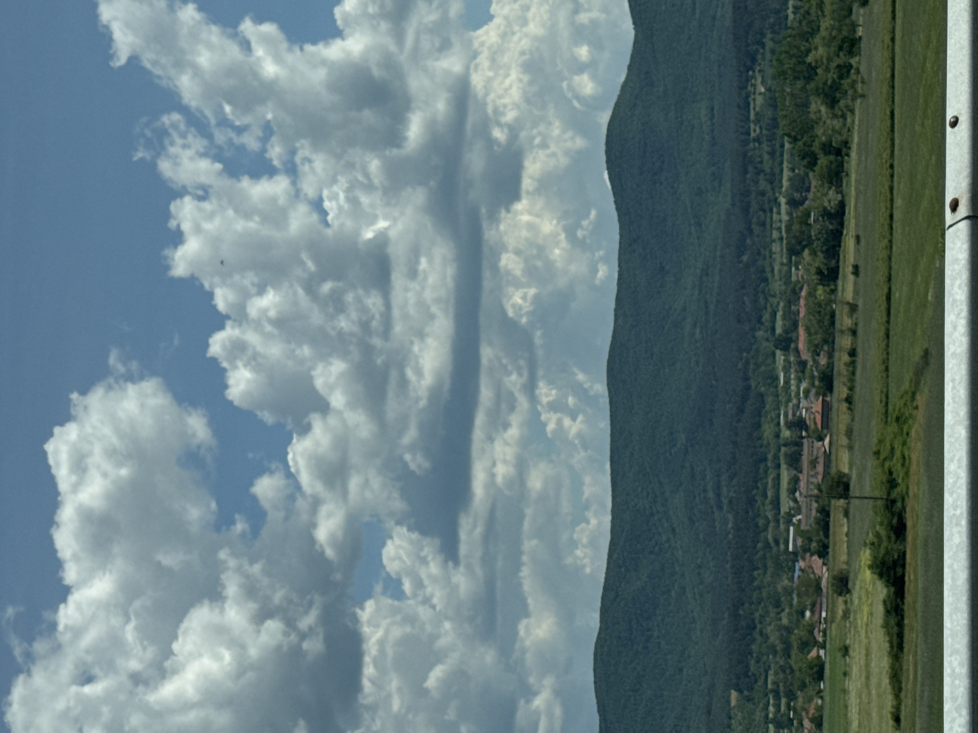 A landscape photograph taken in an unknown location presents a scene of a mountain range bordering a clear blue sky with fluffy white clouds. To the right of the frame, a strip of green land is visible, potentially a field or meadow, adjacent to the mountain's edge. The clouds vary in size and density, some scattered across the sky while others form dense formations. The overall tone of the photograph is serene and peaceful, capturing a scenic view of nature.